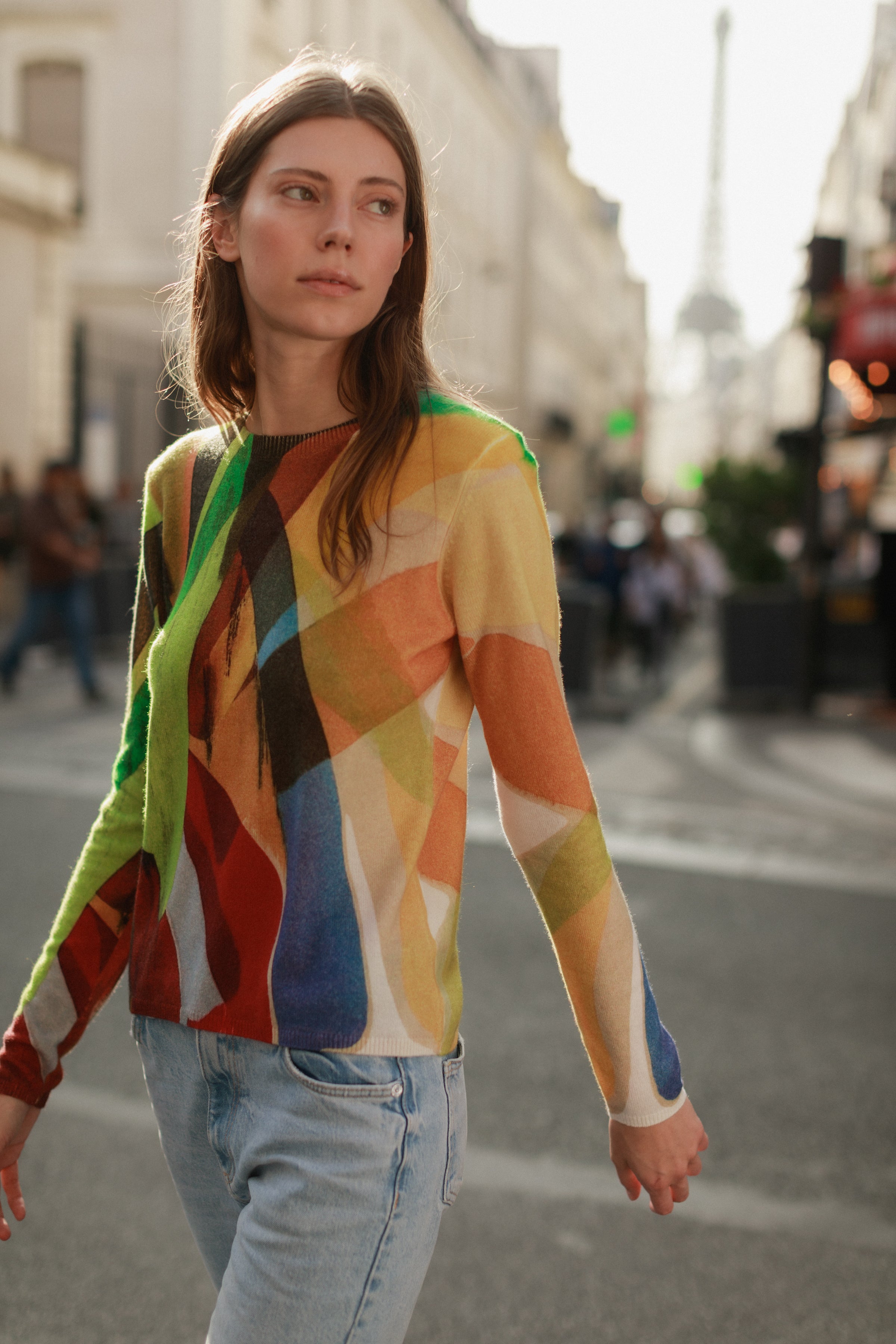 Woman walking on a city street wearing a Catherine Gee Crewneck Cashmere Sweater - Stella Print features a vibrant, abstract pattern with various colors including green, red, yellow, blue, and black paired with light blue jeans. The background shows a blurred cityscape with buildings and the Eiffel-like Tower in the distance, indicating the location is likely Paris.