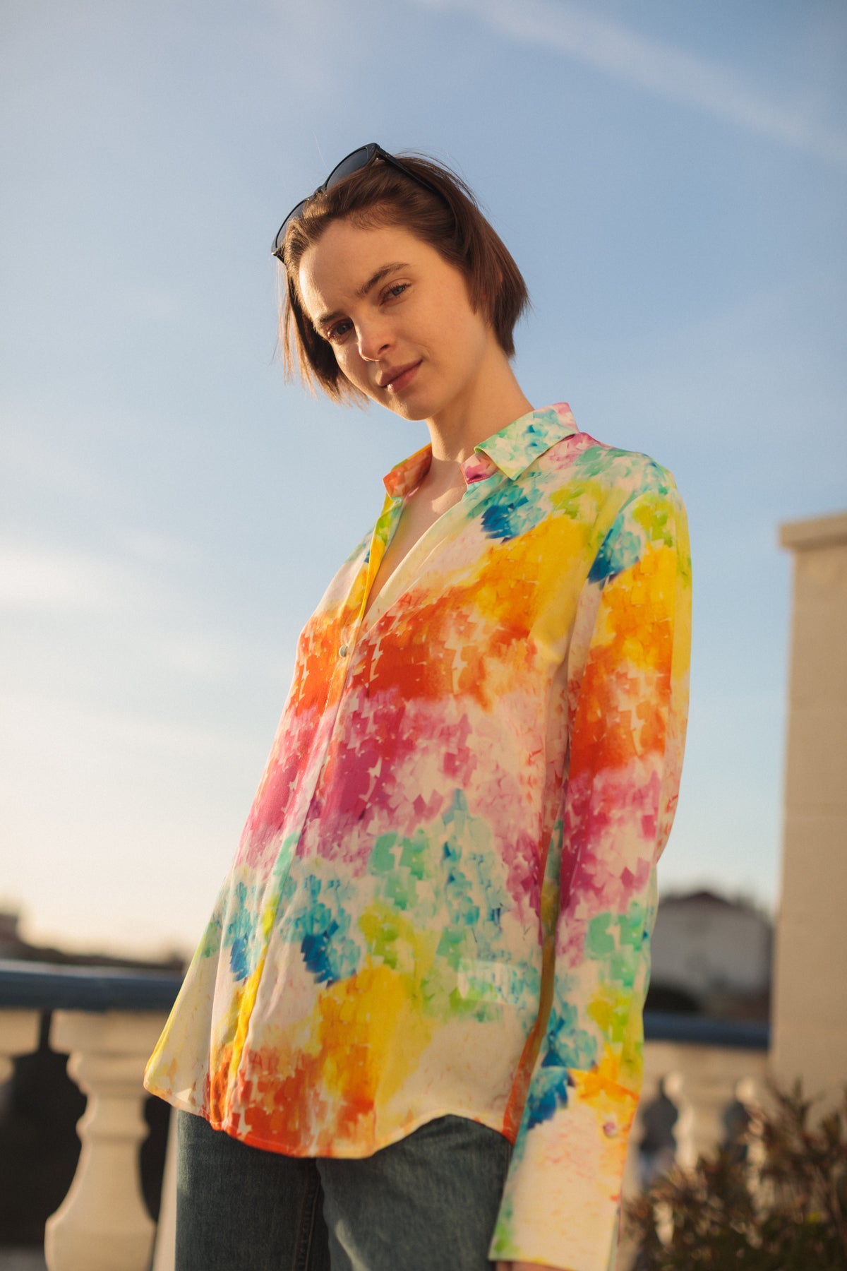 A woman standing outdoors with a clear sky in the background wearing a Catherine Gee Daria French Cuff Silk Rainbow Moodboard blouse features a vibrant, multicolored pattern with splashes of yellow, pink, blue, green, and orange, resembling a watercolor painting. It has a classic collar and buttons down the front. The colorful and artistic design of the blouse makes it visually striking and fashionable.