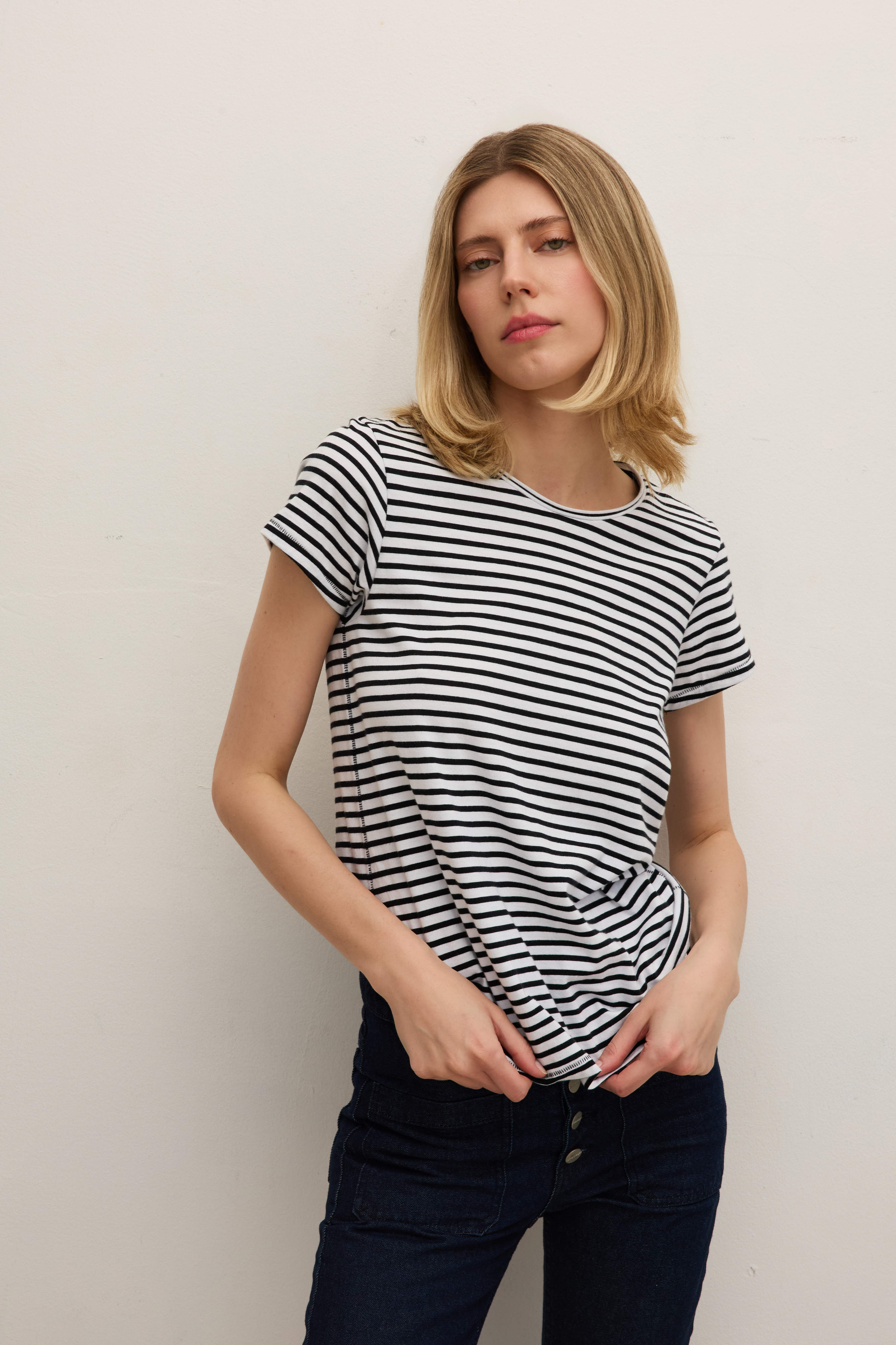 A woman is wearing a black and white striped t-shirt by Catherine Gee paired with dark jeans, standing against a plain white background with her hands resting on her hips. The T-shirt has short sleeves and a round neckline.