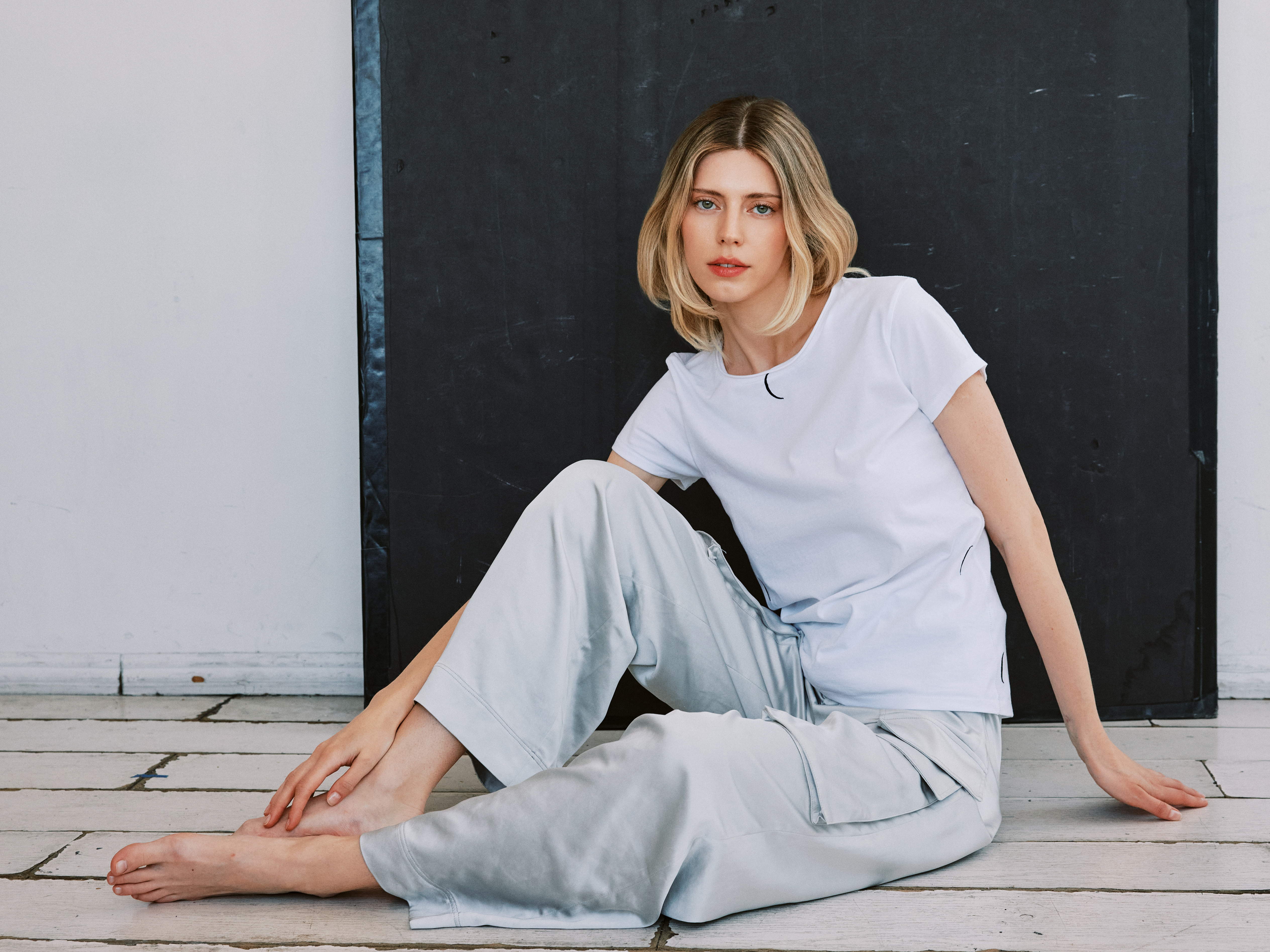 A woman sitting on the wooden floor against a dark background, wearing a white Catherine Gee Embroidered Cotton Crescent T-Shirt paired with light-colored pants features a small crescent moon embroidery near the neckline. The relaxed pose and minimalist style highlight the casual and comfortable nature of the clothing.