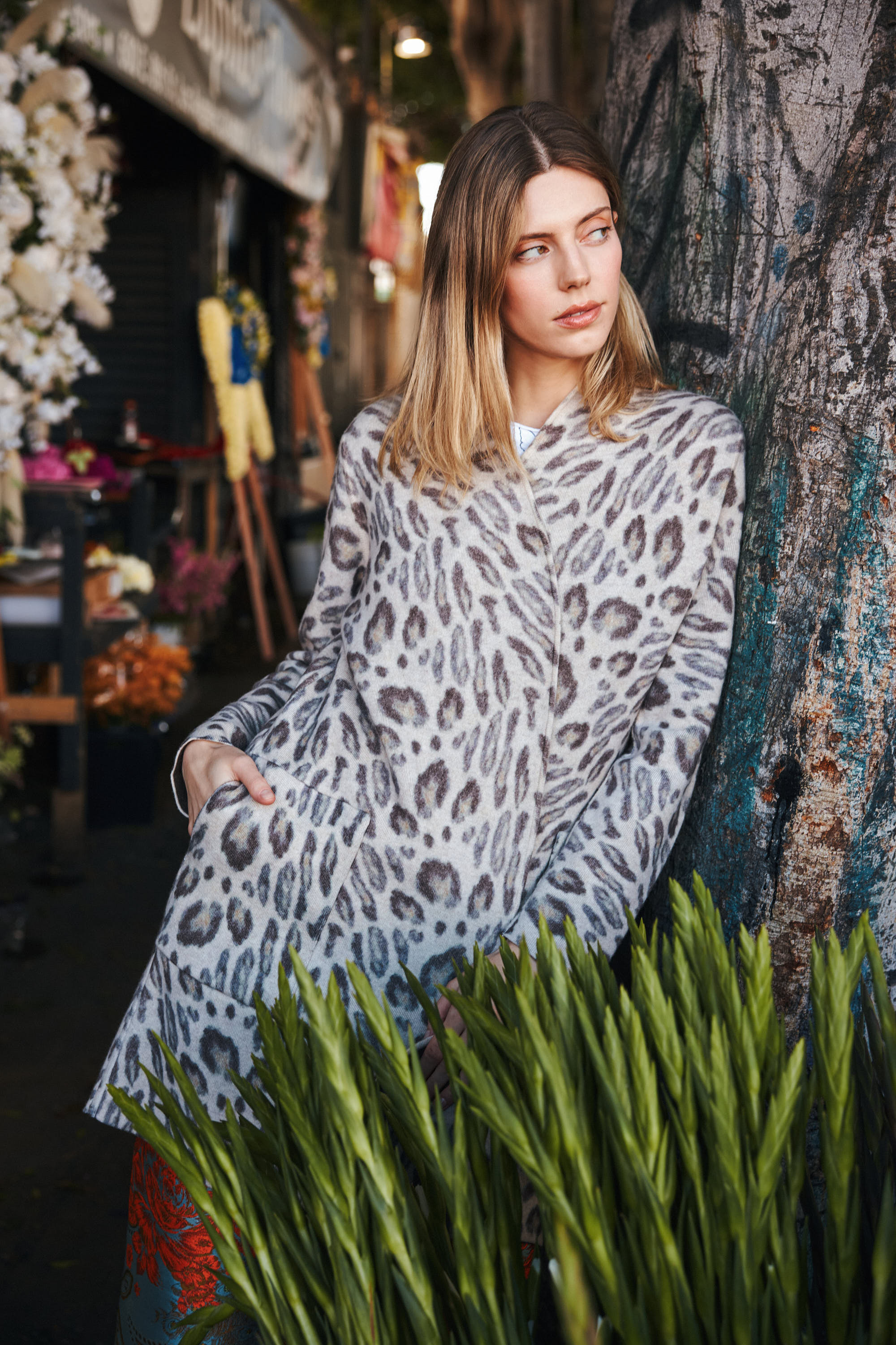 A woman that one hand in the coat pocket leaning against a tree, wearing a Catherine Gee Double Face Cashmere Reversible New Leopard coat features a sophisticated leopard print design, adding a touch of luxury and style to the outfit. The setting appears to be a market, with various plants and flowers in the background highlighting the coat's elegant and versatile design, perfect for making a fashion statement.