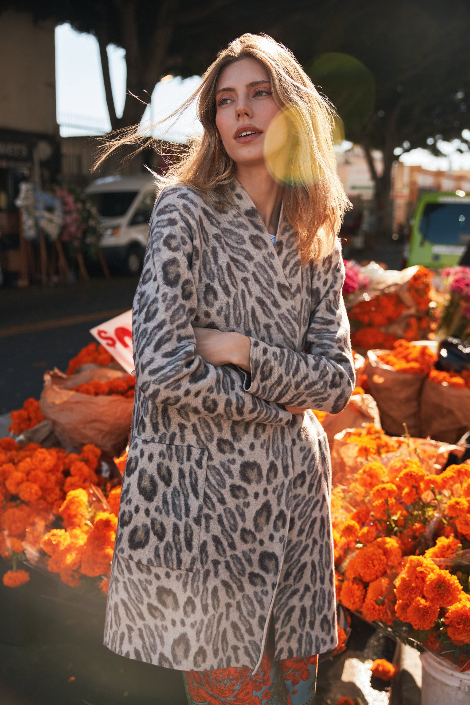 A woman wearing a Catherine Gee Double Face Cashmere Reversible New Leopard coat features a stylish leopard print pattern and appears to be made of high-quality cashmere material. She stands with her arms crossed in front of a vibrant display of orange marigold flowers, adding a colorful and lively background to the scene, enhancing the casual yet chic appeal of the coat.