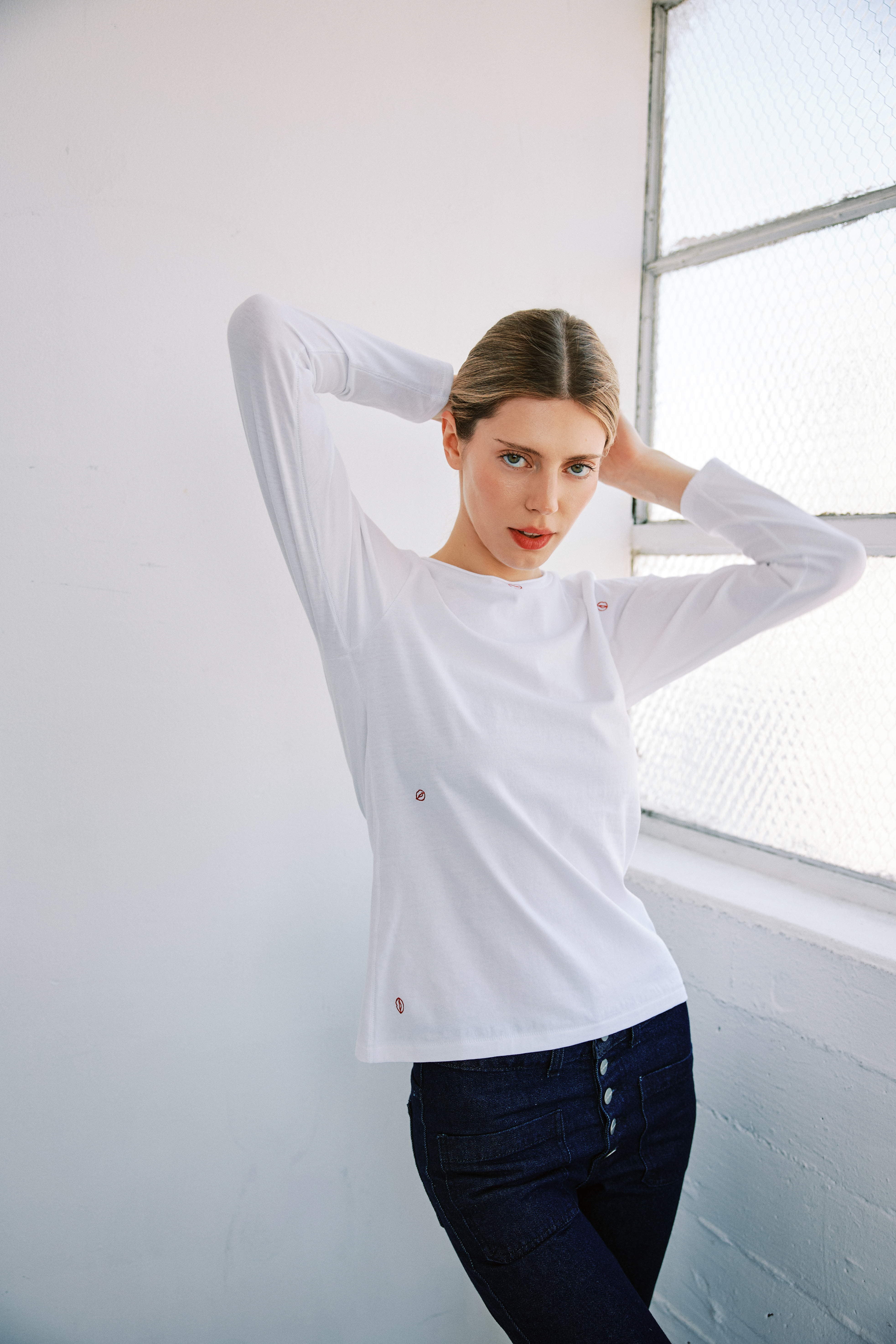 A woman posing with her arms raised next to a window with wire mesh wearing a Catherine Gee Embroidered Cotton Long Sleeve Bisou white t-shirt, which features small embroidered lips near the neckline and sides, adding a charming detail. The setting appears to be a bright, minimalist room, emphasizing the clean and stylish look of the t-shirt. Perfect choice for a comfortable and fashionable outfit.