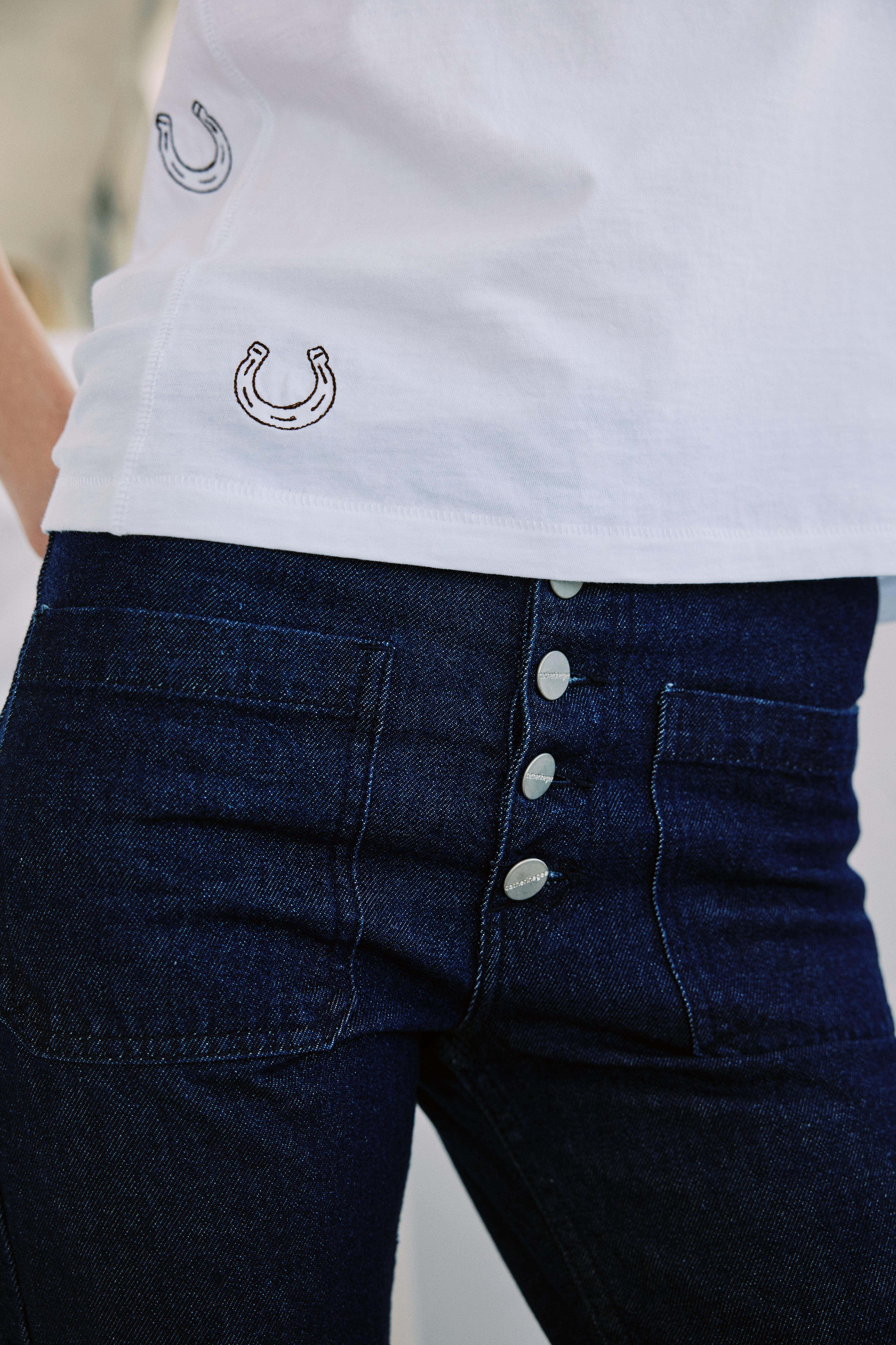A woman wearing a white Catherine Gee Embroidered Cotton Horseshoe T-Shirt paired with dark blue high-waisted denim jeans with a button fly closure, consisting of four visible metallic buttons. The t-shirt features small, delicate horseshoe embroidery scattered near the hem, adding a touch of charm. This combination highlights the t-shirt's detailed embroidery and the jeans' stylish design, perfect for a casual yet fashionable look.