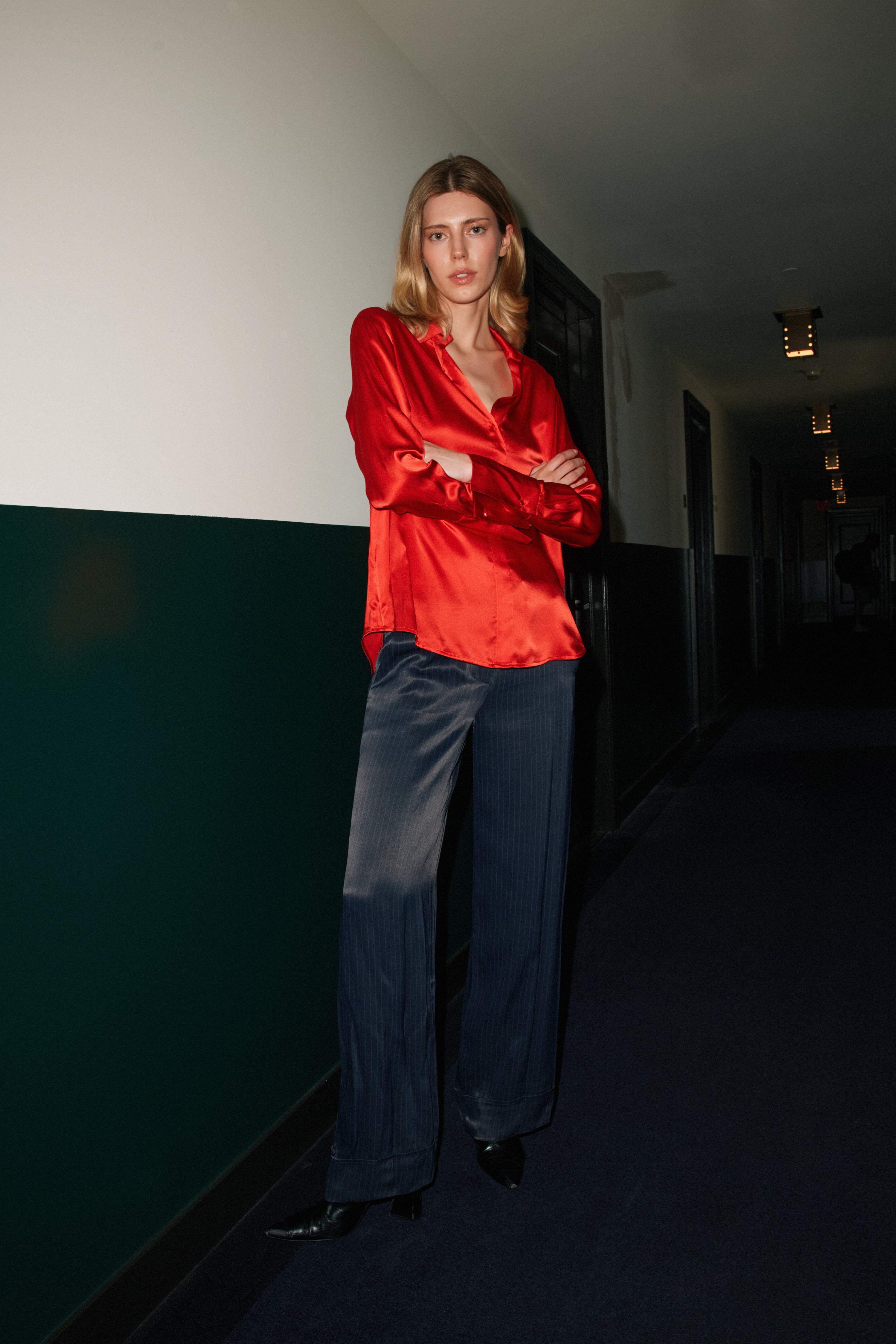 A woman standing in a hallway with arms crossed, wearing a Catherine Gee Daria French Cuff Silk-High Risk Red blouse features vibrant red silk blouse and dark wide-leg pants. The hallway has a green and white wall with a dark blue carpet with dim lighting, creating a moody atmosphere.