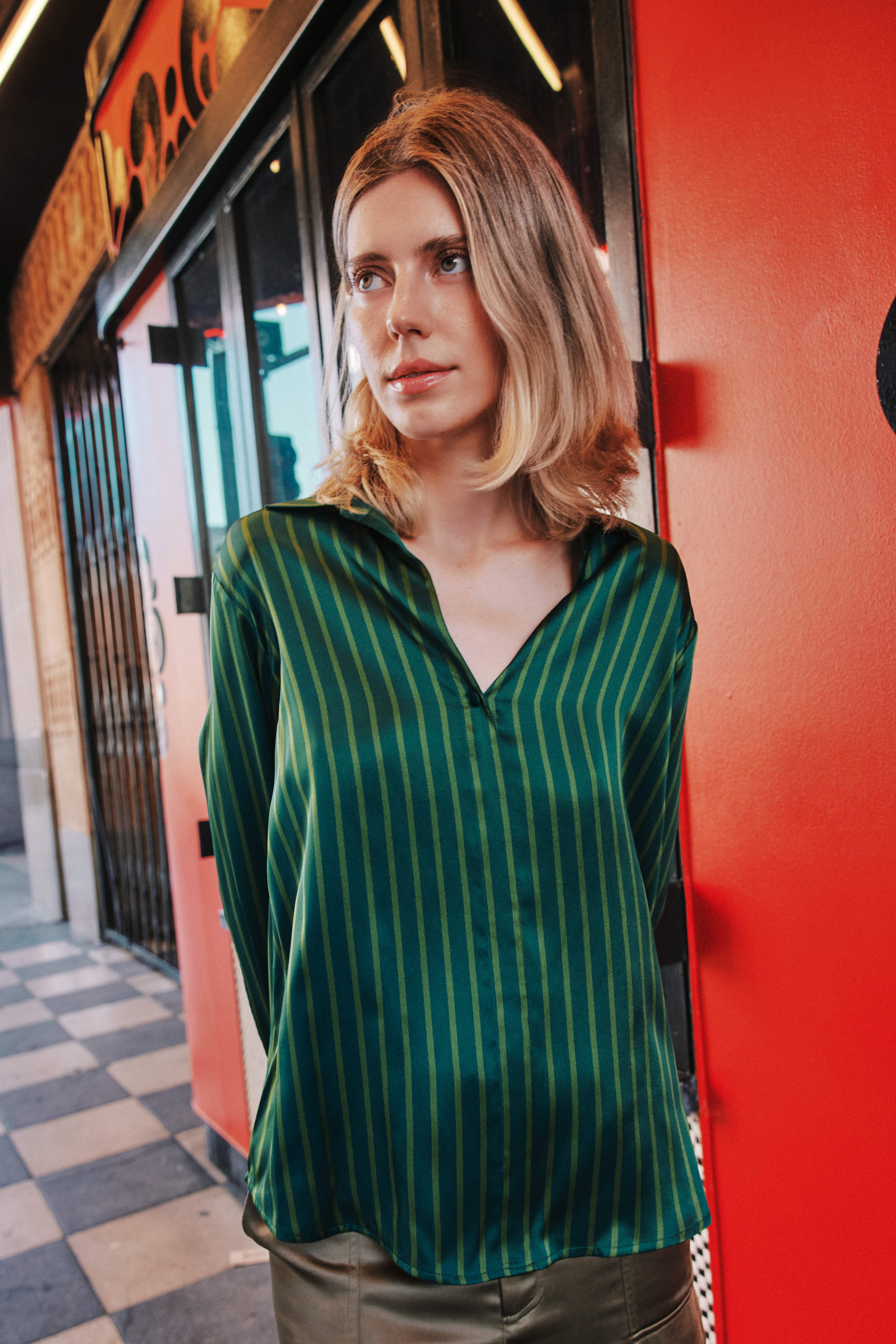 A woman wearing a Catherine Gee Daria French Cuff Silk-Green Stripes blouse features a V-neck and long sleeves. The background includes a red wall and a checkered floor, suggesting an urban setting.