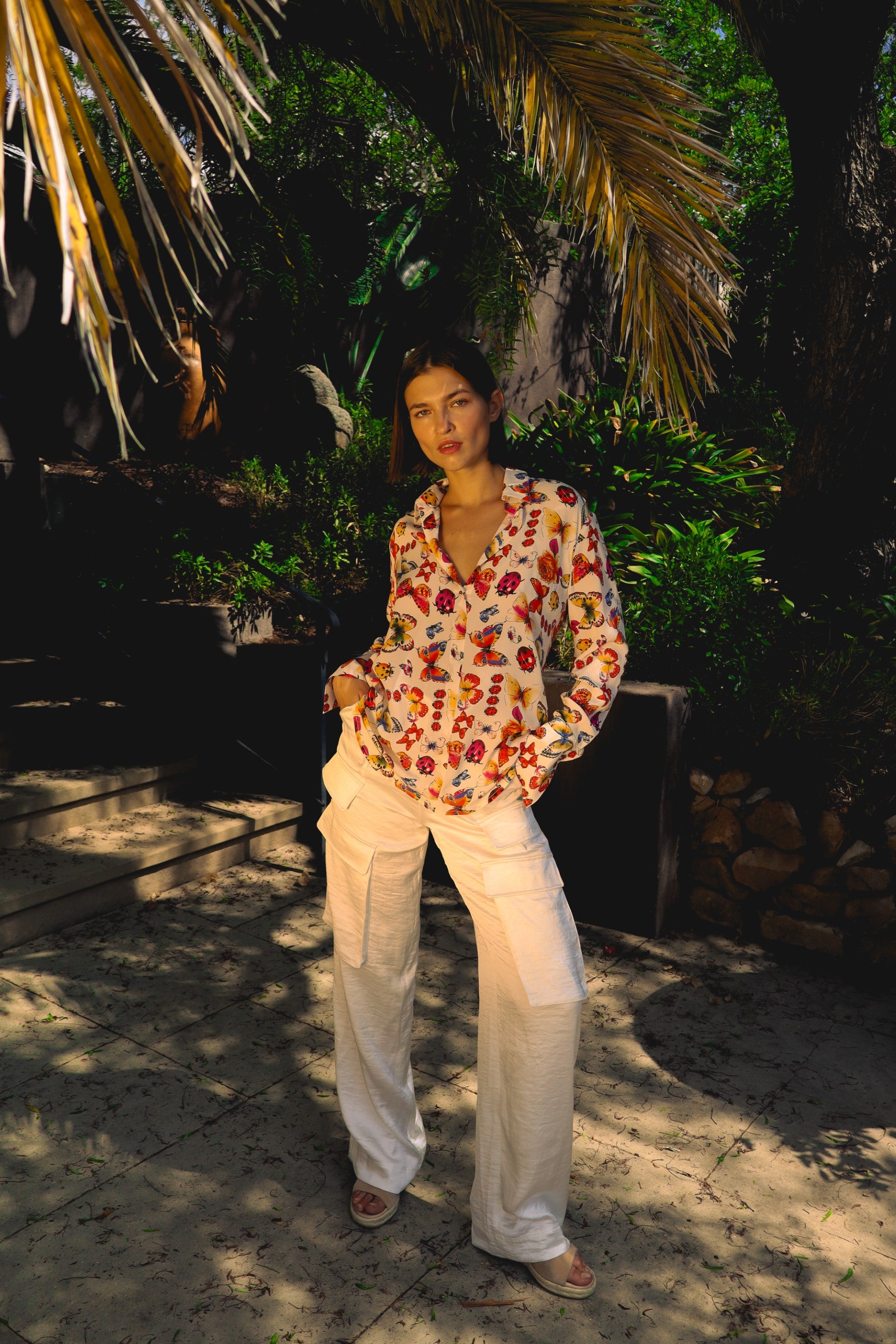 A woman standing outdoors in a shaded area with lush greenery and palm fronds in the background wearing a Catherine Gee Daria French Cuff Silk Blouse with a ladybug print, paired with white pants and sandals. The setting appears to be a garden or park with stone steps and a tiled pathway highlighting the blouse's vibrant pattern and the relaxed, stylish outfit.