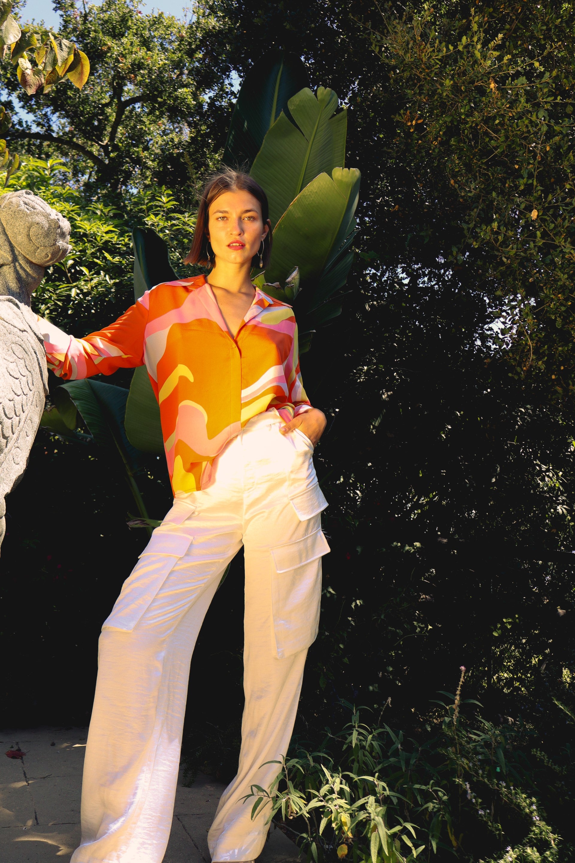 Women standing outdoors in a lush, green environment wearing a vibrant Catherine Gee Daria French Cuff Silk Sherbet Blouse, characterized by its bold, colorful pattern in shades of orange, pink, yellow, and white. The blouse has a relaxed fit and is paired with white, high-waisted pants featuring multiple pockets. She is posing with one hand on a stone sculpture and the other in a pocket, creating a stylish and confident look.