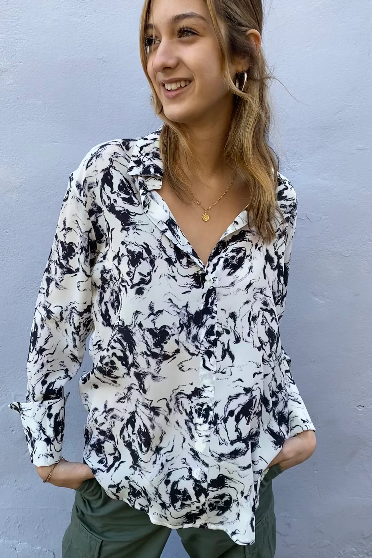 A woman is wearing a Catherine Gee Daria French Cuff Silk black and white abstract blouse that features a loose fit with a V-neckline and long sleeves with French cuffs. She has her hands in the pockets of her green pants and is accessorized with a gold necklace and hoop earrings on a plain light-colored wall background.