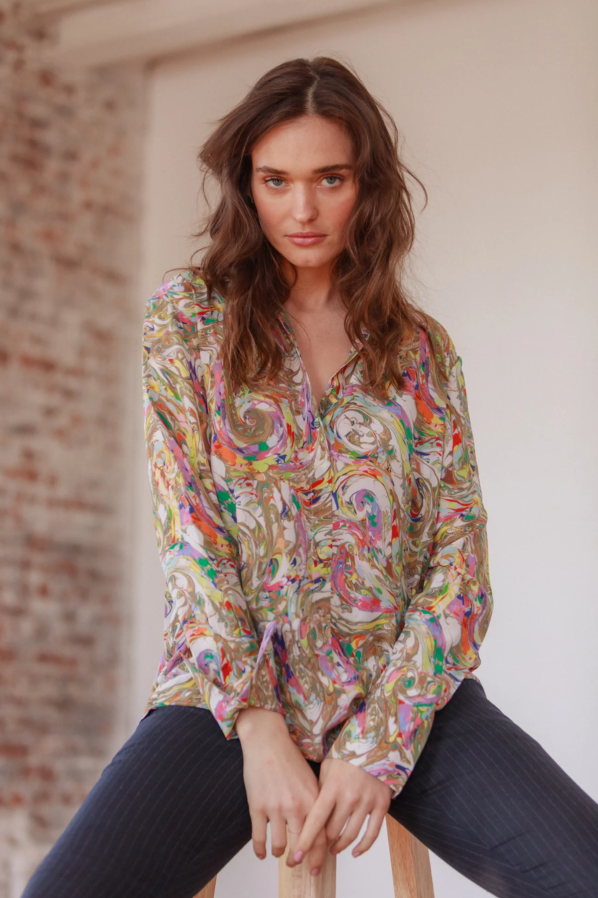 A woman wearing a Catherine Gee Daria French Cuff Silk Paint Swirl blouse features a vibrant and intricate pattern with swirling colors, including shades of gold, red, purple, blue, and green. The fabric appears to be lightweight and silky, giving it a luxurious and elegant look. The blouse has a V-neckline and French cuffs, adding to its sophisticated design. She is seated on a stool, with the background showing a partially visible brick wall and a white wall, creating a casual yet stylish setting.