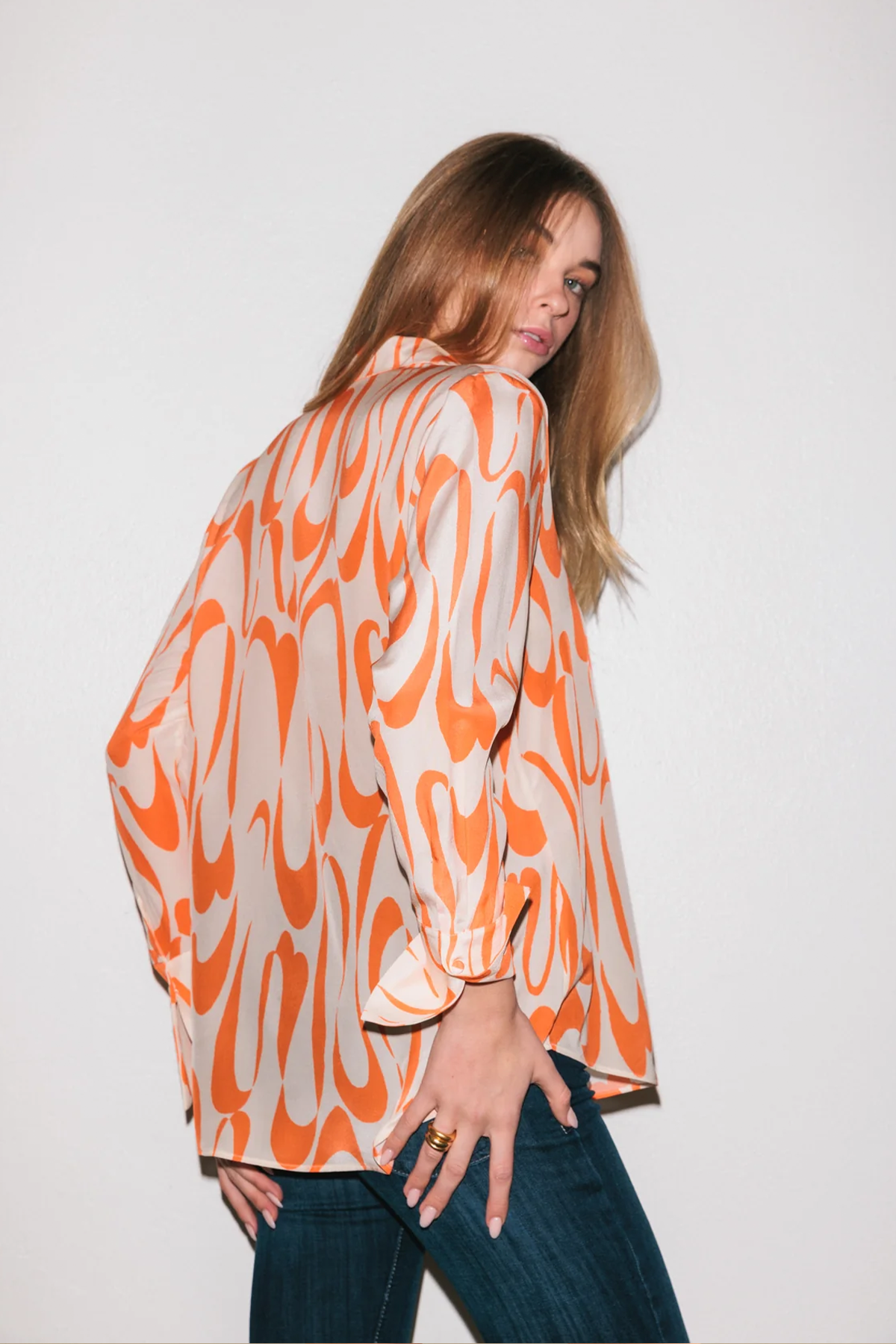 A woman wearing the Catherine Gee Daria French Cuff Silk Valencia blouse features a vibrant orange and cream abstract pattern, with a loose fit and French cuffs. The silky texture and bold design make it a standout piece. She also wears dark blue jeans, which complement the blouse's striking colors, creating a fashionable and elegant outfit.