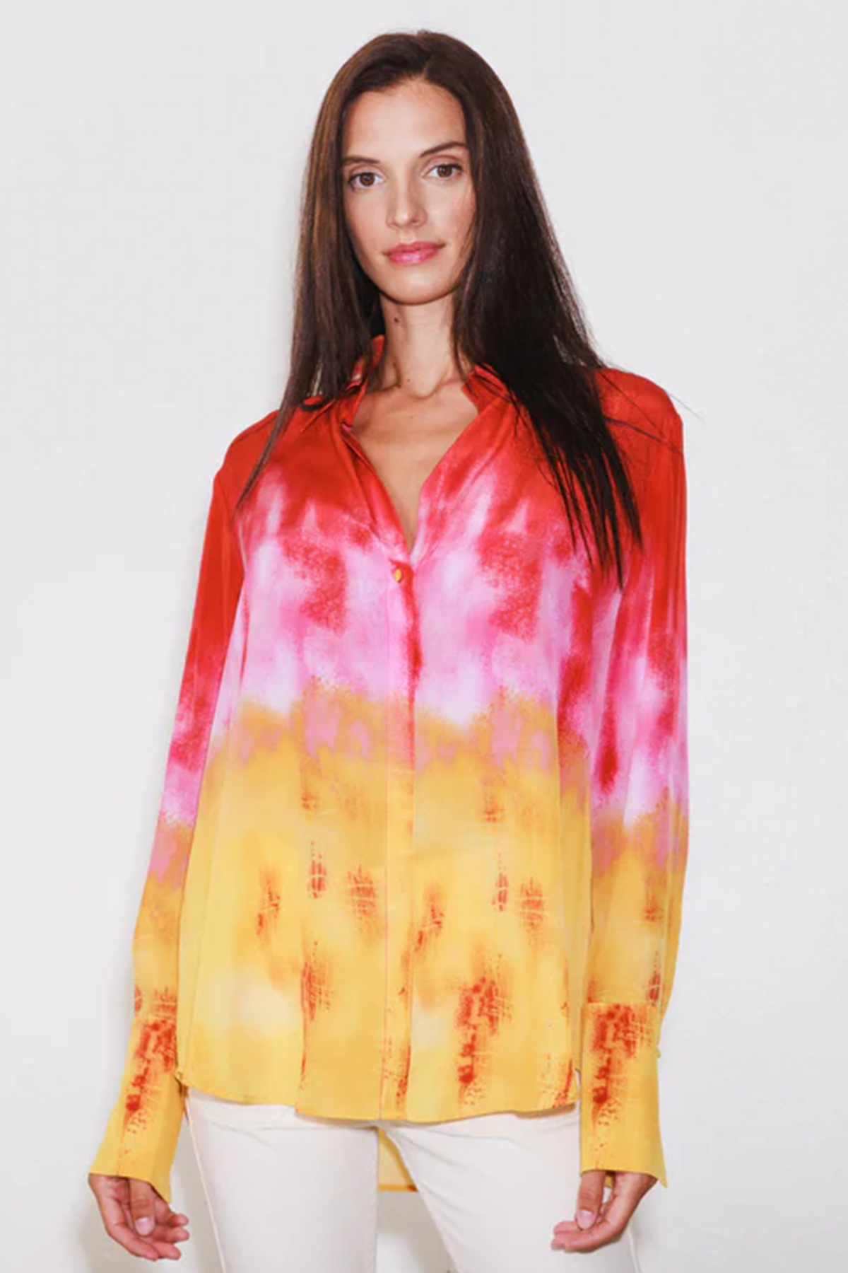 Catherine Gee Daria French Cuff Silk Santa Barbara Sunset blouse features a vibrant gradient design, transitioning from red at the top to pink in the middle, and yellow at the bottom. The fabric appears to be silk, giving it a smooth and shiny texture and has a button-down front and long sleeves with French cuffs. She wears the blouse paired with white pants, highlighting the blouse's colorful design.