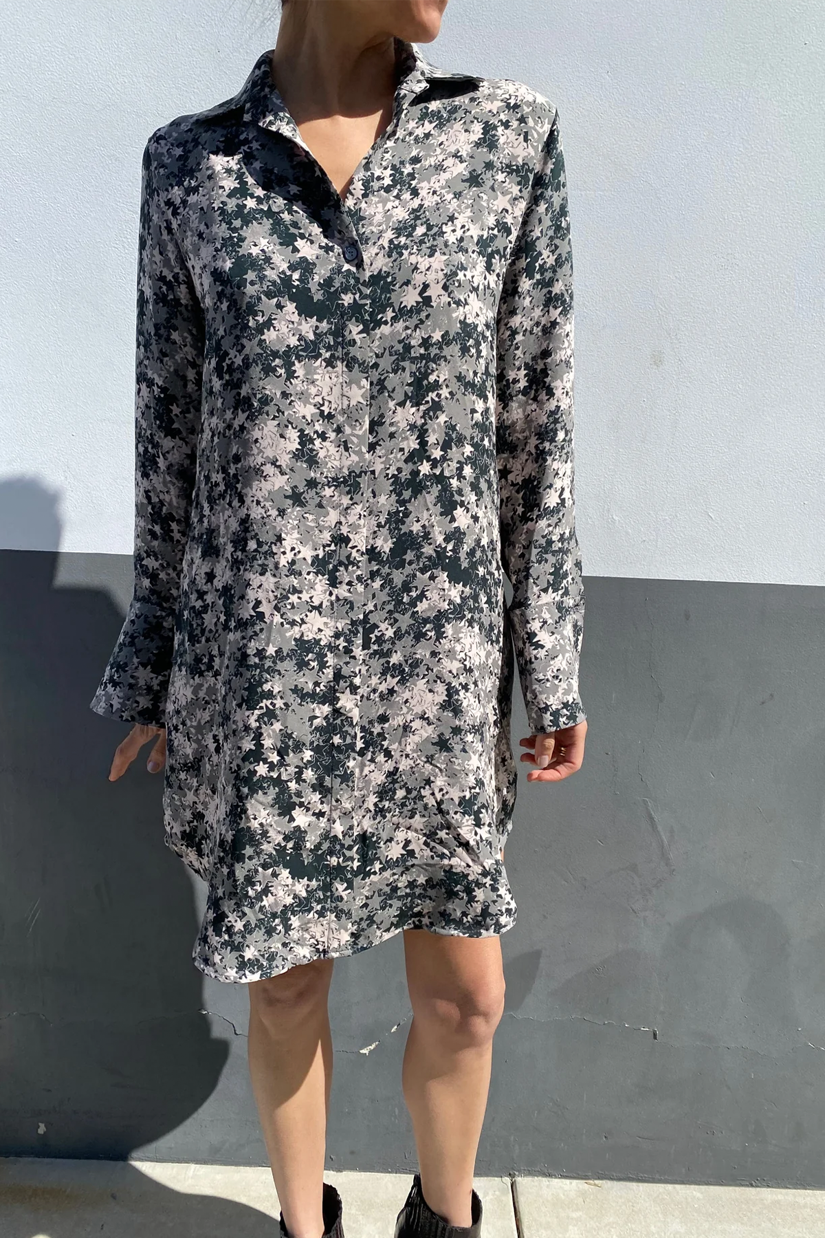 A woman wearing a Catherine Gee Daria Silk Shirt Starshower Dress. This elegant dress features a collar and long sleeves, with a button-down front. The fabric showcases a beautiful star-shower pattern with scattered star-like shapes in black and white and has a loose, comfortable fit that falls above the knees, offering a blend of casual comfort and stylish sophistication. The background is a plain wall with a two-tone color scheme, enhancing the dress's visual appeal.