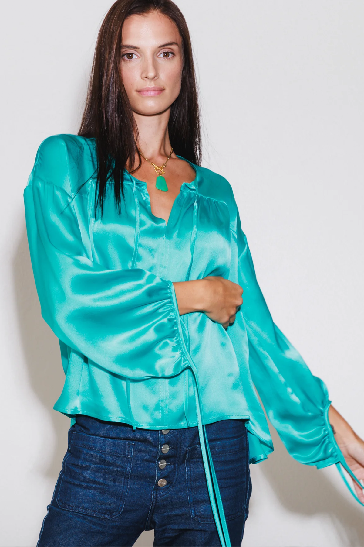 A woman wearing the Catherine Gee Frances Van Gogh Green Blouse. This vibrant green blouse is crafted from luxurious silk charmeuse, featuring a relaxed, open silhouette with a V-neckline and long, loose sleeves gathered at the cuffs. Paired with high-waisted dark blue jeans and accessorized with a green pendant necklace, the ensemble highlights the blouse's elegant and stylish design, perfect for day-to-day wear.