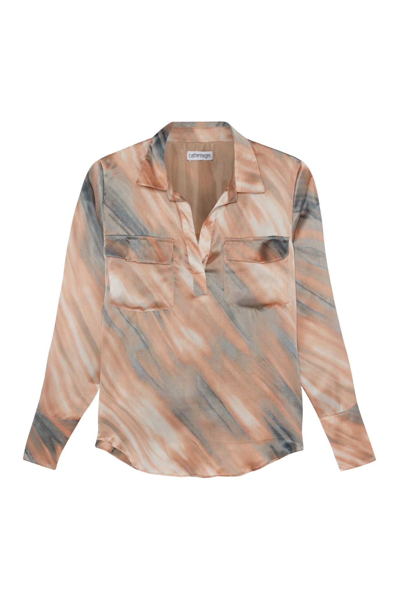 Catherine Gee Kaia Dune Blouse, a long-sleeved, button-up shirt with a collar. The blouse features a unique, abstract pattern in shades of peach, beige, and gray, resembling a marbled or dune-like design. It has two chest pockets and a relaxed fit, making it a stylish and versatile piece suitable for various occasions. The fabric appears lightweight and flowy, adding to its elegant and comfortable appeal.