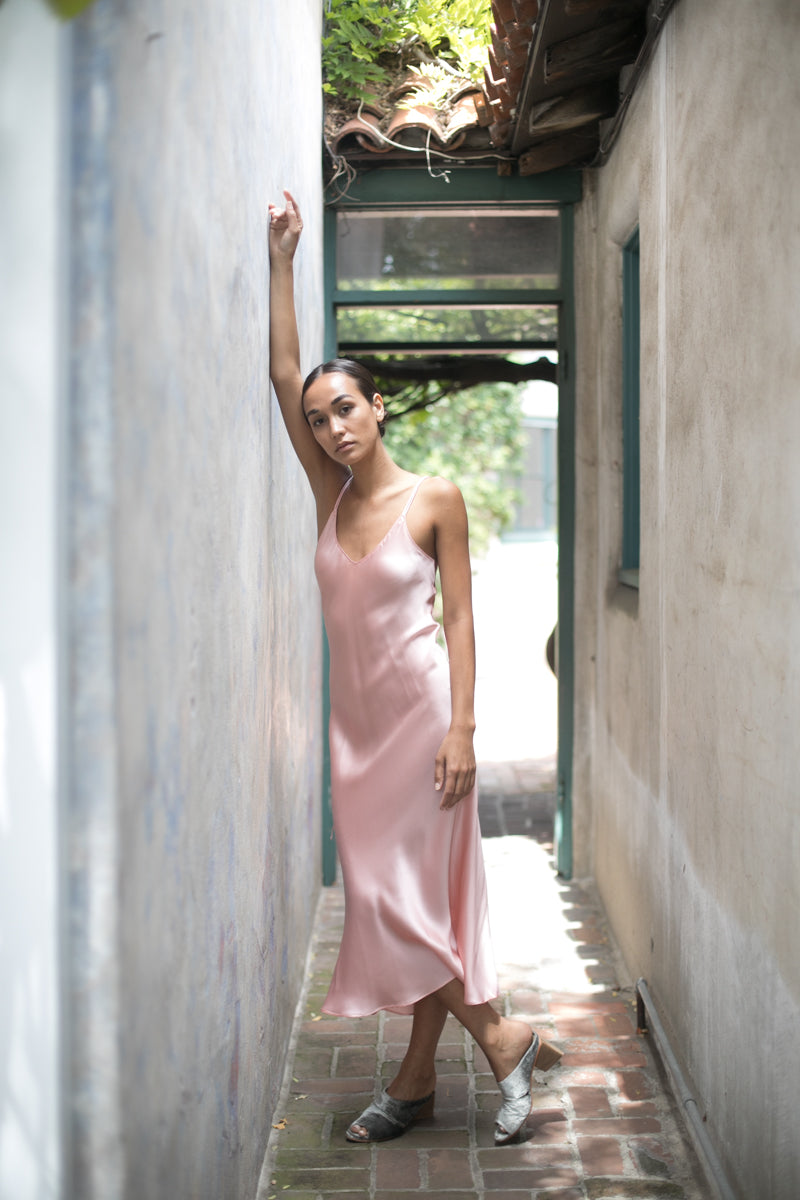 A woman leaning against a wall in a narrow alleyway, one arm raised and the other resting by her side wearing the Catherine Gee Emma Pink Slip Dress, which is a silky light pink slip dress featuring thin straps and a mid-length hemline. The setting is outdoors, with a tiled floor and a partially covered roof overhead, leading to a bright, leafy background. The dress exudes effortless elegance and sophistication.