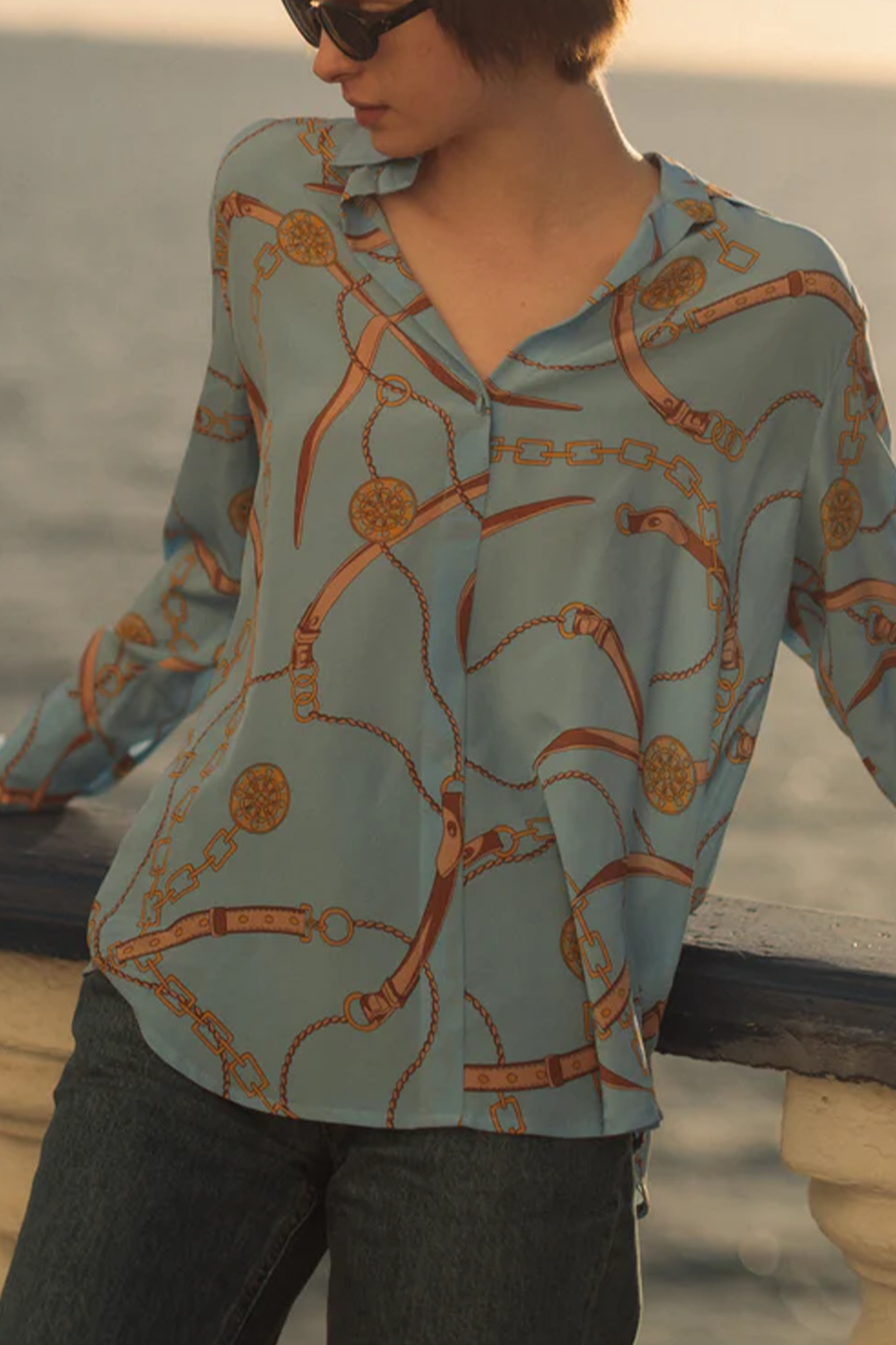A woman wearing a Catherine Gee Daria French Cuff Silk San Telmo blouse features a light blue base color with an intricate pattern of gold chains and buckles. The fabric appears to be soft and flowing, indicative of high-quality silk, and has a relaxed fit with a v-neckline and long sleeves, making it both stylish and elegant. The background is near water on the sea, adding to the serene and sophisticated ambiance.