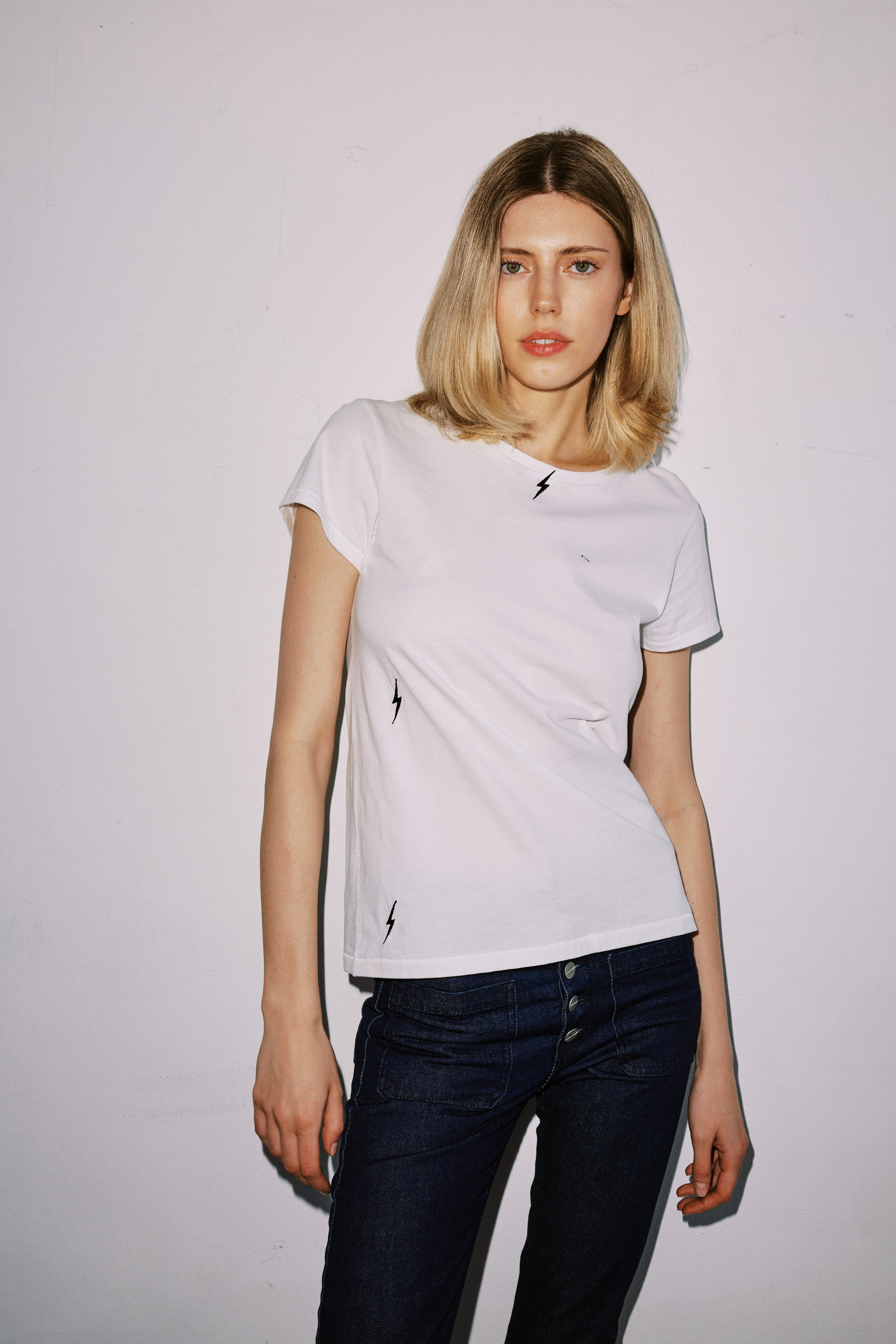 A woman wearing a white Catherine Gee Embroidered Cotton White Bolt t-shirt features small black lightning bolt embroidery on the chest and lower side paired with dark blue jeans. The plain white background emphasizes the t-shirt's minimalist yet stylish design, making it a versatile piece for casual wear.