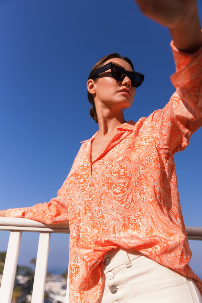 A woman wearing a Catherine Gee Daria French Cuff Silk Sorbet Swirl blouse features a vibrant orange and white swirl pattern, creating a dynamic and eye-catching look. Made from luxurious silk, it has a relaxed fit and long sleeves with French cuffs. She is standing outdoors with a clear blue sky, with one arm resting on a railing and the other extended, possibly taking a selfie. The blouse's lightweight material and stylish design make it both fashionable and comfortable.