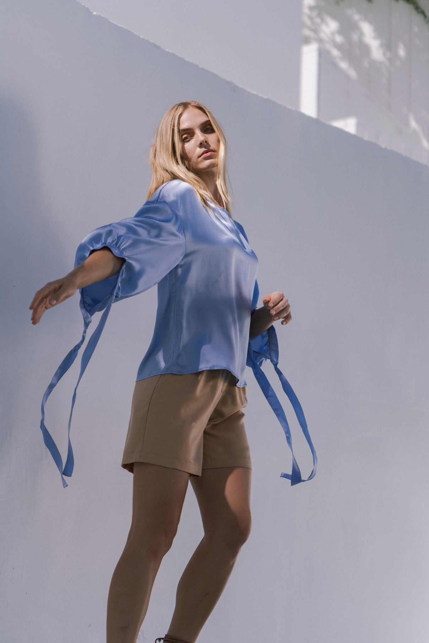 A woman is wearing a sky-blue Catherine Gee Constance Blouse with long, flowing ties on the sleeves paired with beige shorts. The person is standing against a light-colored wall with arms slightly extended, creating a dynamic and graceful pose, emphasizing the elegance of the blouse.