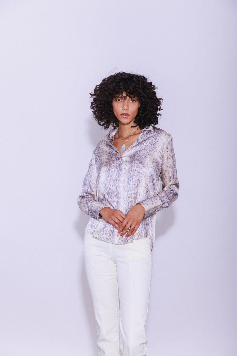 A woman wearing a Catherine Gee Daria French Cuff Silk Sedona Snake blouse features a chic snake print pattern in shades of grey and white. It has a silky texture and a relaxed fit with a button-down front and French cuffs, adding a sophisticated touch paired with white pants. The overall look is stylish and elegant, showcasing the blouse's luxurious fabric and contemporary design.