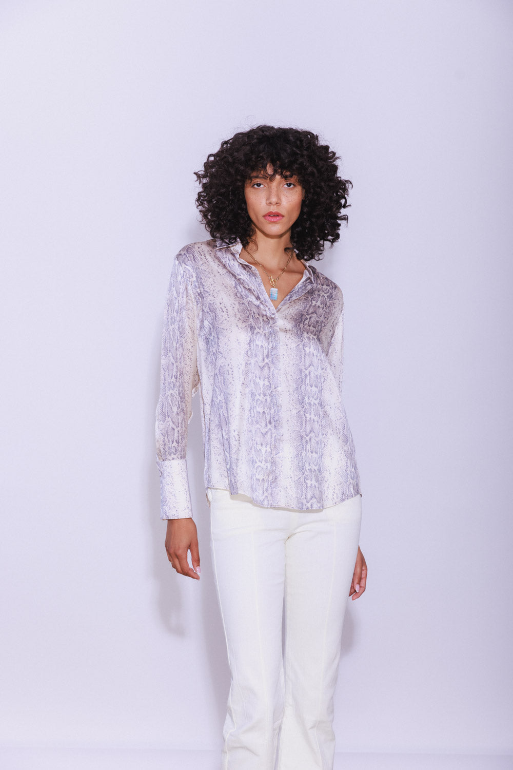 A woman wearing a Catherine Gee Daria French Cuff Silk Sedona Snake blouse features a subtle snake print pattern in shades of grey and white, with a relaxed fit and a V-neckline. The long sleeves end in French cuffs, adding a touch of sophistication paired with white pants, creating a chic and elegant look that highlights the blouse's luxurious silk fabric and stylish design.