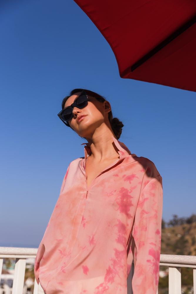 A woman wearing a Catherine Gee Daria French Cuff Silk Pink Ink blouse features a pink color with ink-like patterns, giving it a unique and artistic look. It has a classic collar and is made of a smooth, silky fabric. She is standing outdoors under a red umbrella with a clear blue sky on a sunny day helps the blouse stand out due to its vibrant color and elegant design, making it a stylish and eye-catching piece of clothing.