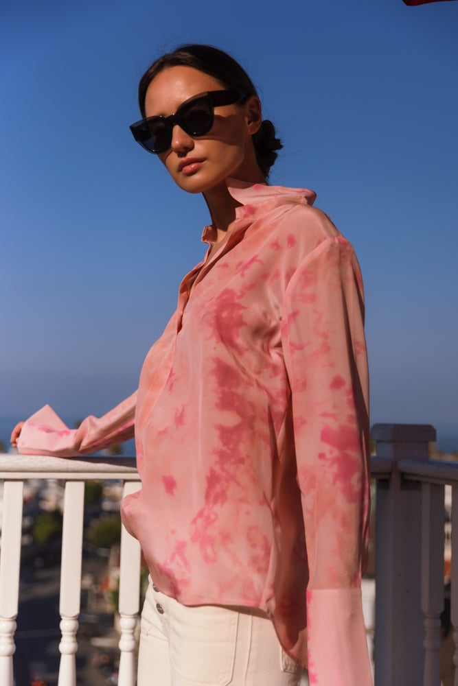 A woman wearing a Catherine Gee Daria French Cuff Silk Pink Ink blouse features a vibrant pink color with a unique ink-like pattern, creating a dynamic and eye-catching design paired with white pants. The fabric appears to be silky and lightweight, providing a smooth and luxurious texture, a classic collar, and long sleeves with French cuffs, adding a touch of sophistication. She is standing on a balcony with a clear blue sky on a bright sunny day complementing the soft and elegant look of the outfit.