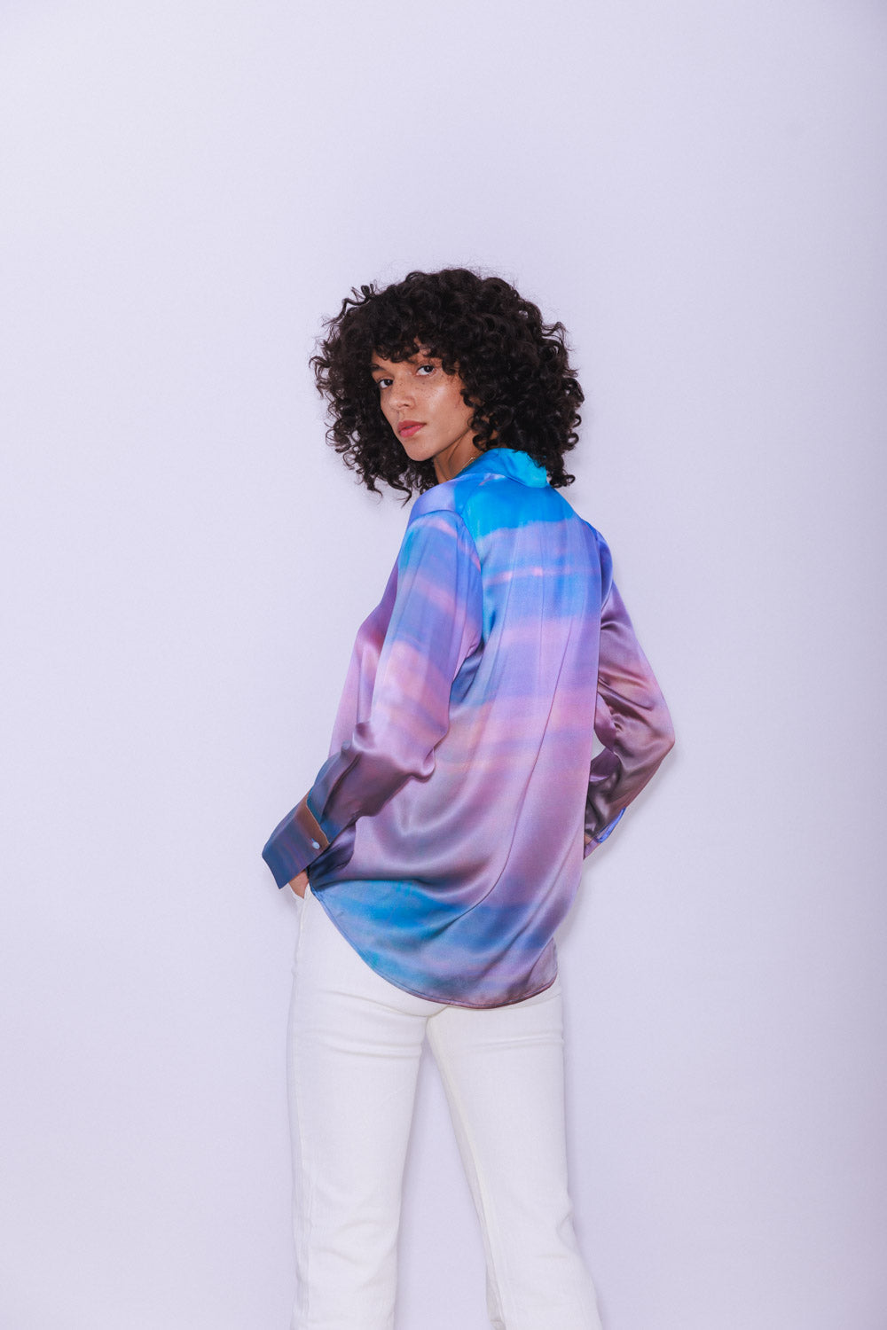 A woman wearing a Catherine Gee Daria French Cuff Silk Peruvian Quartz blouse features a vibrant, multicolored pattern with shades of blue, purple, and pink, resembling a quartz-like design. The blouse has a silky texture and a loose fit, with long sleeves and French cuffs paired with white pants highlighting the unique and colorful design of the blouse, making it interesting for fashion enthusiasts.