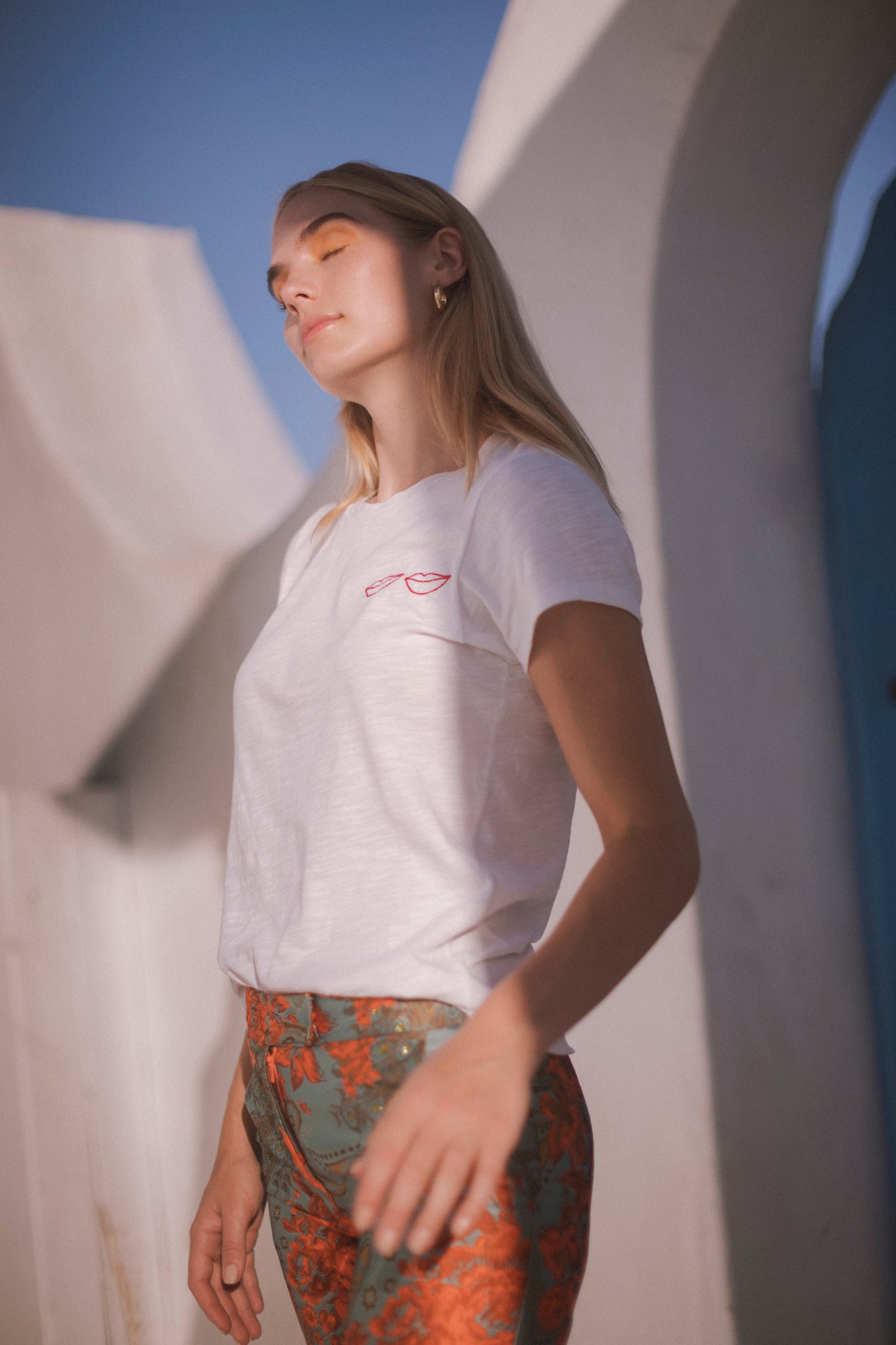 A woman wearing a Catherine Gee Embroidered Cotton Lips white t-shirt features a small, embroidered design of red lips on the left side of the chest paired with high-waisted pants featuring a green and orange floral pattern. The backdrop includes white architectural elements and a clear blue sky, suggesting a bright, outdoor setting that combines casual elegance with a playful touch.