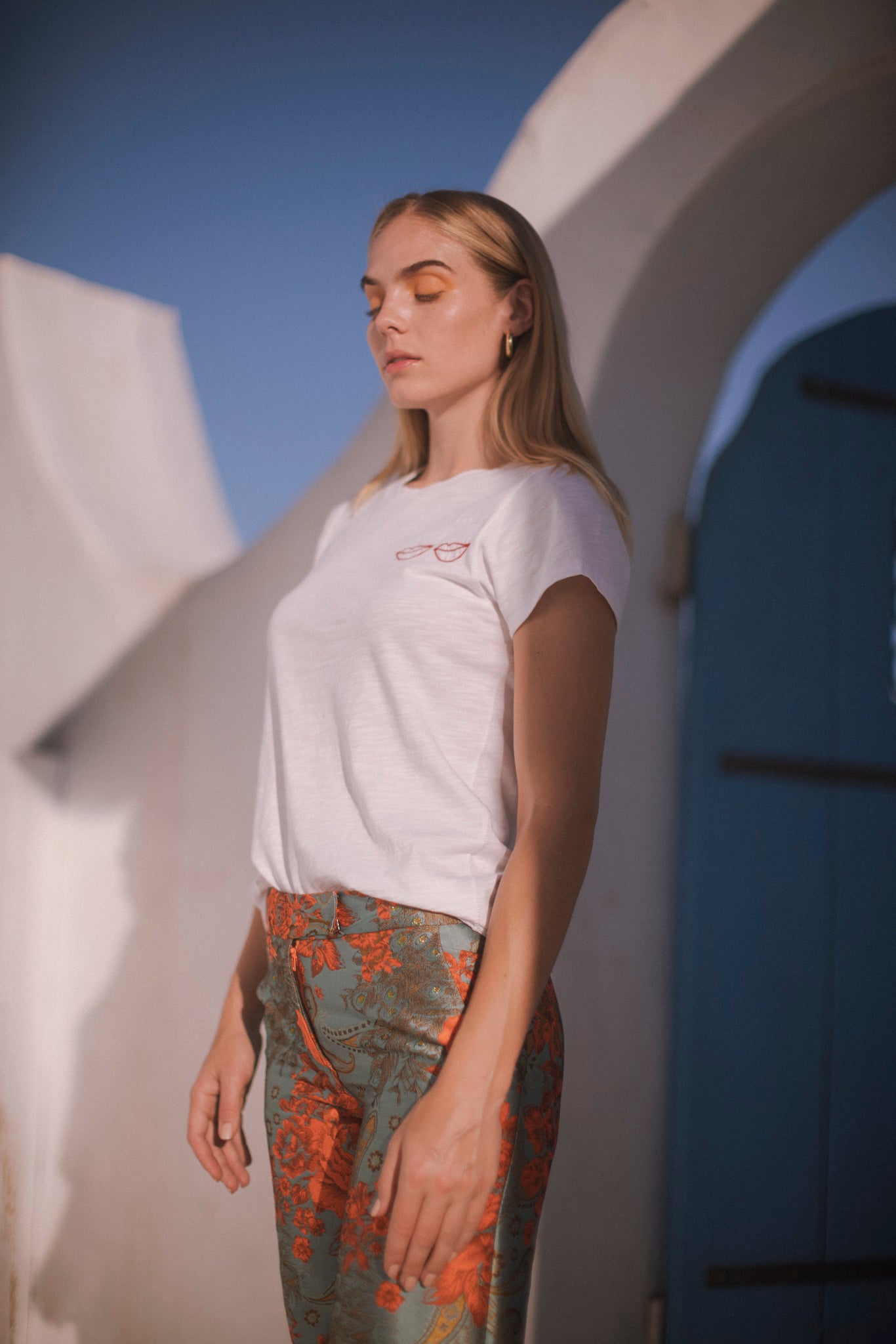 A woman wearing Catherine Gee Jacquard Stella Blue/Orange pants features an intricate jacquard pattern with a combination of blue and orange hues, creating a visually striking and elegant design paired with a white t-shirt, and the background includes architectural elements with white walls and a blue door, suggesting a Mediterranean or coastal setting. The overall composition highlights the stylish and sophisticated nature of the pants.