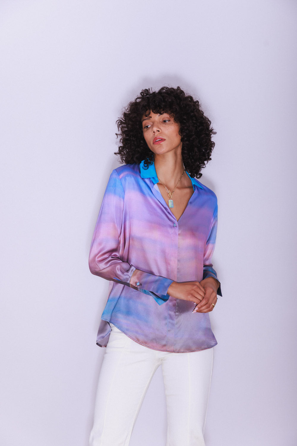 A woman wearing a Catherine Gee Daria French Cuff Silk Peruvian Quartz blouse features a captivating gradient pattern with shades of blue, purple, and pink, creating a visually stunning effect. It has a button-down front and elegant French cuffs paired with white pants and a pendant necklace, adding to the sophisticated look. The plain, light-colored background enhances the vibrant colors of the blouse.