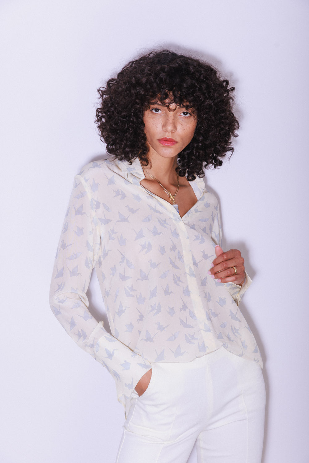 A woman with one hand in the pocket and the other hand holding the blouse wearing a Catherine Gee Daria French Cuff Silk Neutral Crane blouse with a light color with a subtle crane bird pattern creates a delicate and elegant look. It features long sleeves with French cuffs, a straight collar, and a relaxed fit paired with white pants. The plain background emphasizes the details and design of the blouse.