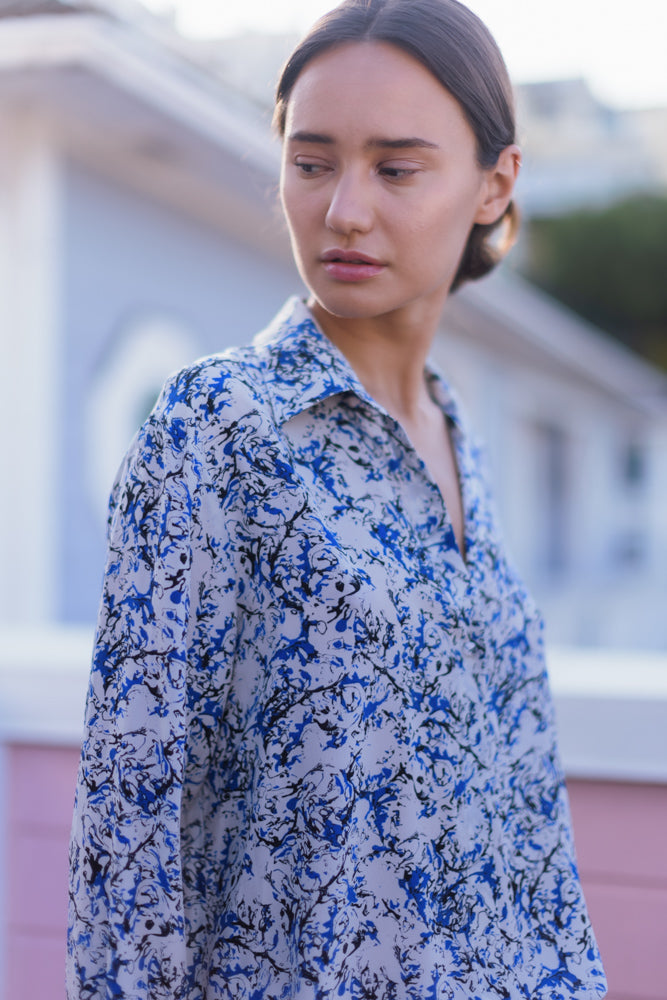 A woman wearing a Catherine Gee Daria French Cuff Silk Santorini blouse features a blue and white abstract pattern with a button-down front and a classic collar. The fabric appears lightweight and silky, giving it a luxurious and elegant look.