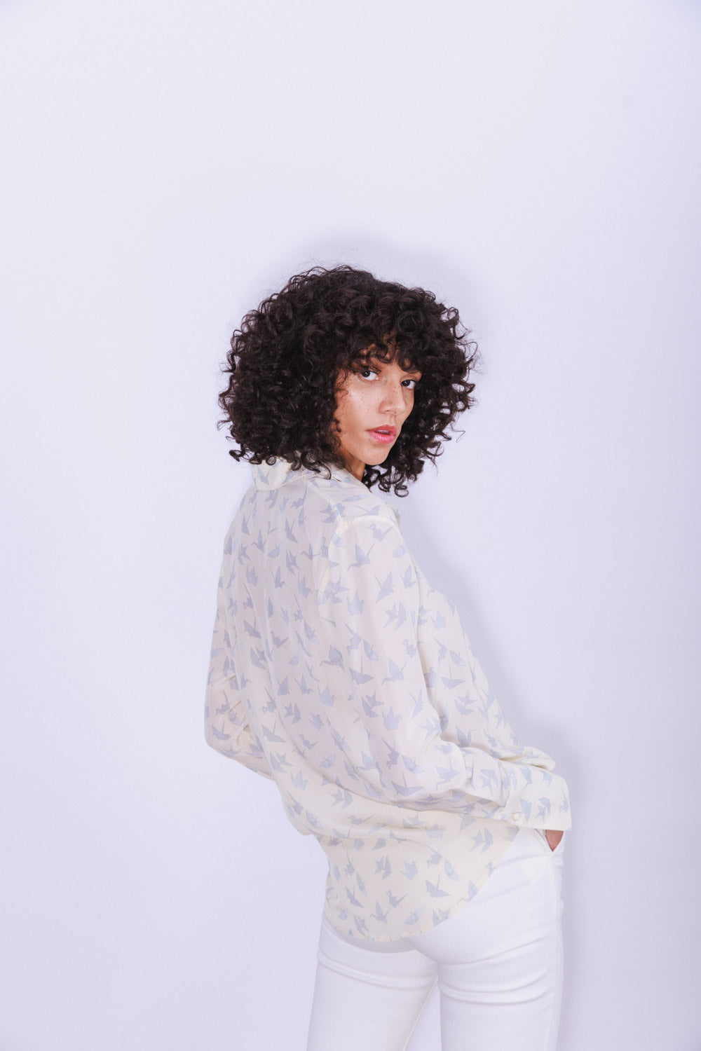 A woman standing with one hand in a pocket, wearing a Catherine Gee Daria French Cuff Silk Neutral Crane blouse in light-colored with a subtle crane bird pattern appears to be made of a lightweight, flowy silk material paired with white pants. The blouse has a relaxed fit and features French cuffs. The overall look is elegant and sophisticated, suitable for both casual and semi-formal occasions.