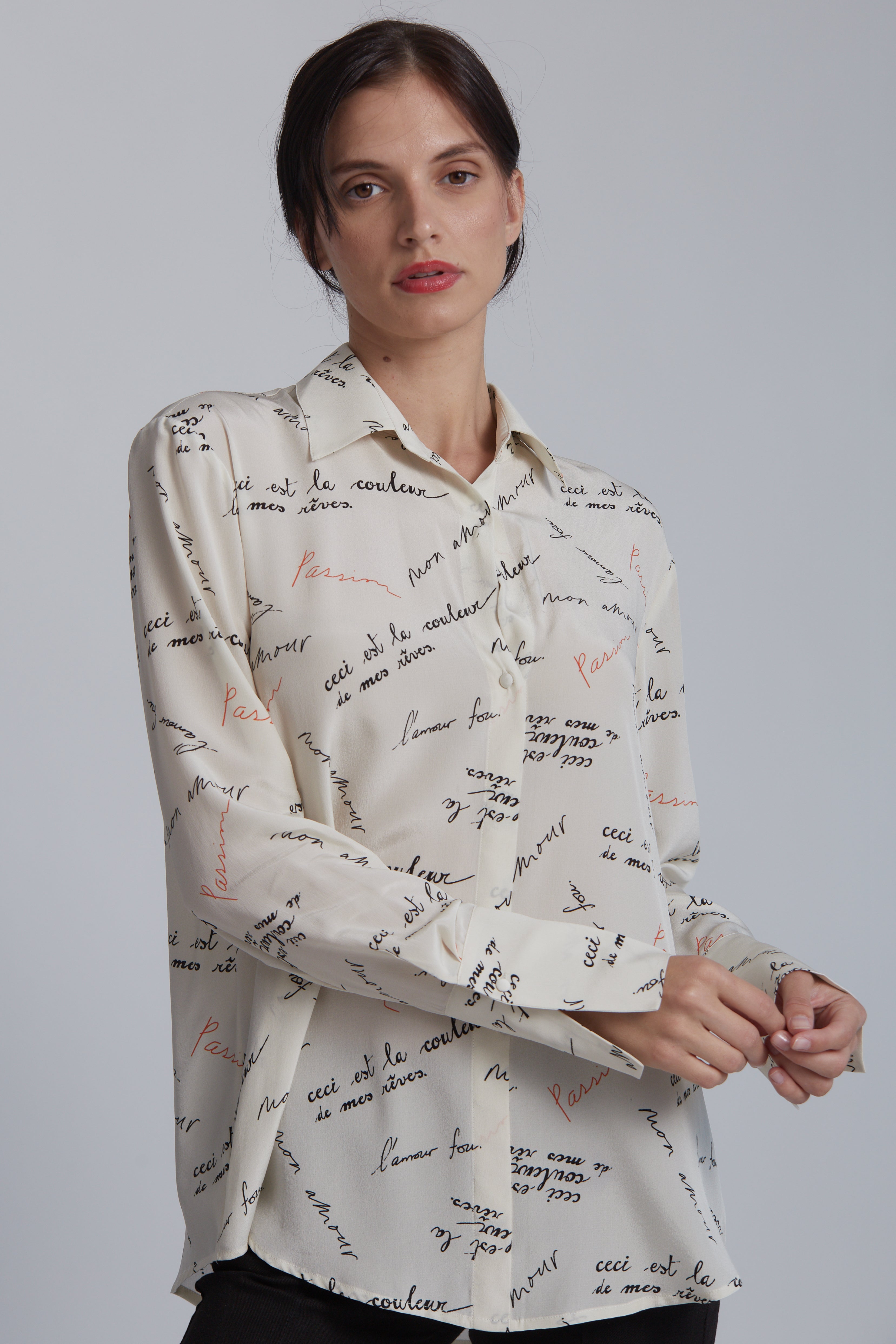 A woman wearing a Catherine Gee Daria French Cuff Silk Passion Print blouse made of silk features a print with various phrases in French, written in different fonts and colors, predominantly black and red. The blouse has a classic collar and button-down front, with long sleeves ending in French cuffs on a plain white background, which helps to highlight the details of the blouse.