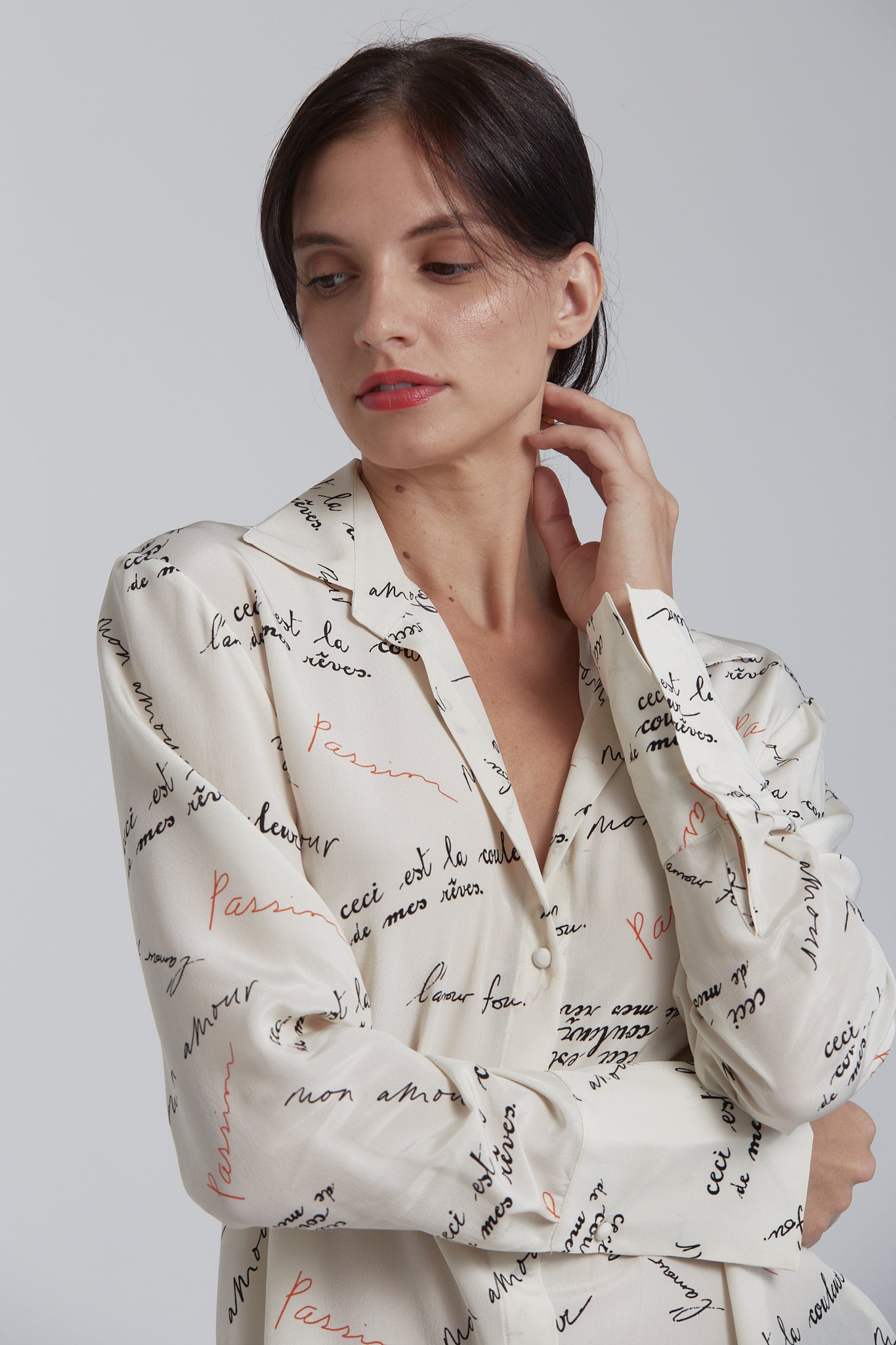 A woman wearing a Catherine Gee Daria French Cuff Silk Passion Print blouse featuring elegant cream-colored and adorned with a pattern of handwritten text in black and red, featuring French phrases like "Ceci est la couleur de mes rêves" and "Passions." The blouse boasts a classic collar, button-down front, and long sleeves with French cuffs. The silk fabric gives it a smooth and luxurious appearance, making it a sophisticated and fashionable choice perfect for a stylish and unique refined wardrobe.