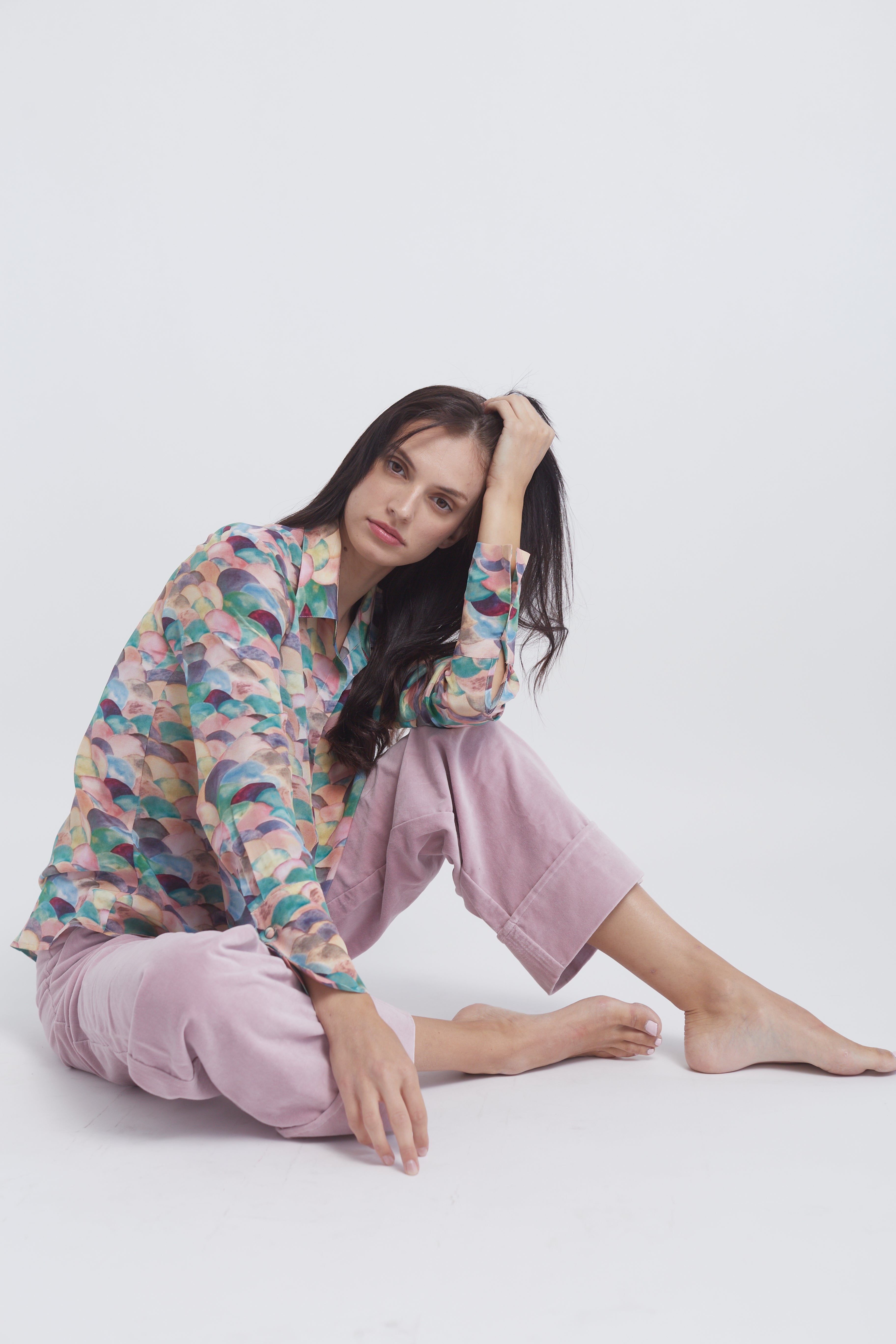A woman seated on the floor with one leg bent and the other extended wearing a colorful, patterned silk blouse with a design that includes various shades of pink, blue, green, and purple, the Catherine Gee Daria French Cuff Silk Blouse Lima paired with light pink pants that are rolled up at the cuffs.