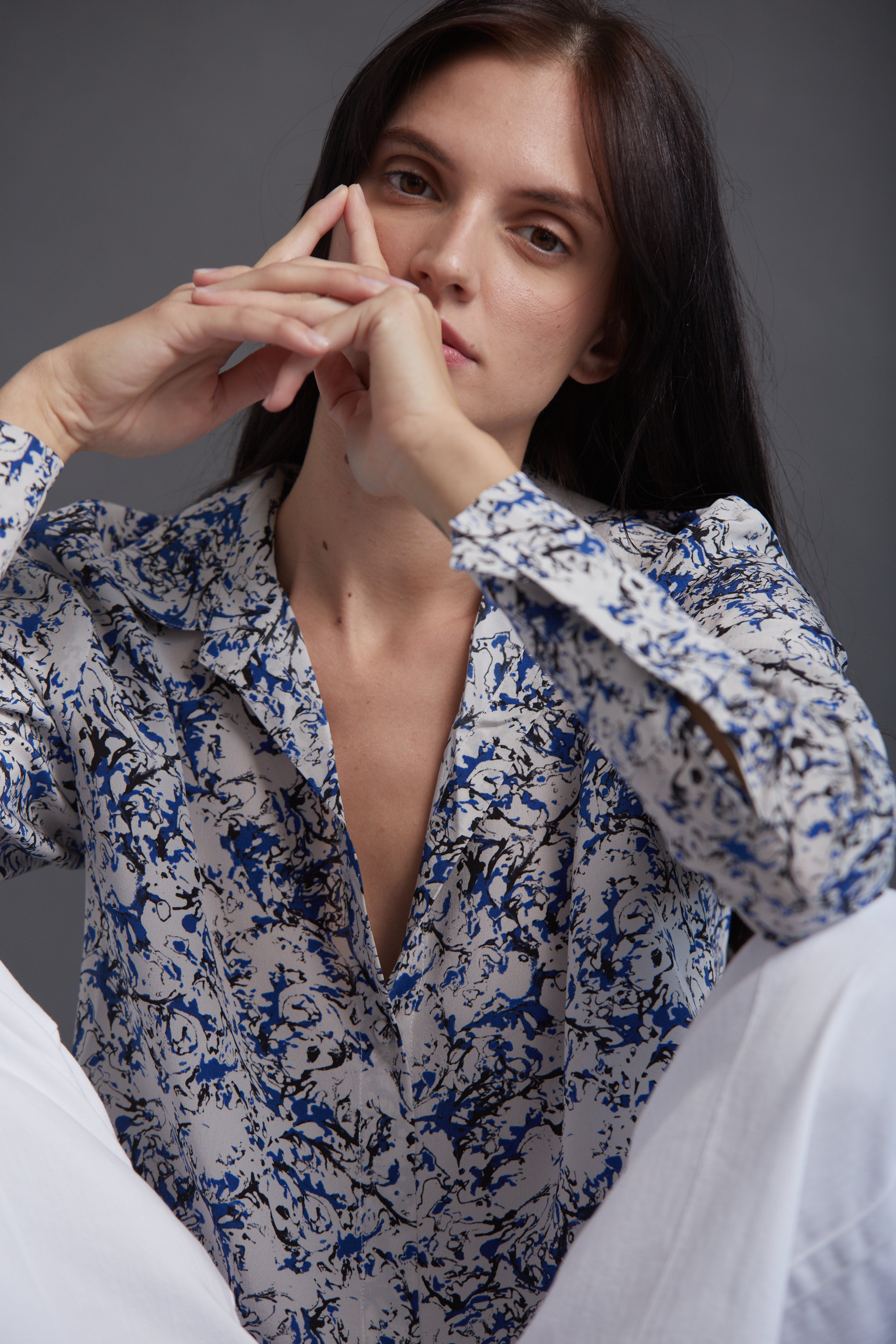 A woman wearing a Catherine Gee Daria French Cuff Silk Santorini blouse features a detailed blue and white abstract pattern with a deep V-neckline and French cuffs. The fabric appears to be lightweight and silky, contributing to an elegant and sophisticated look. She is seated, with arms partially raised, highlighting the blouse's design and texture.