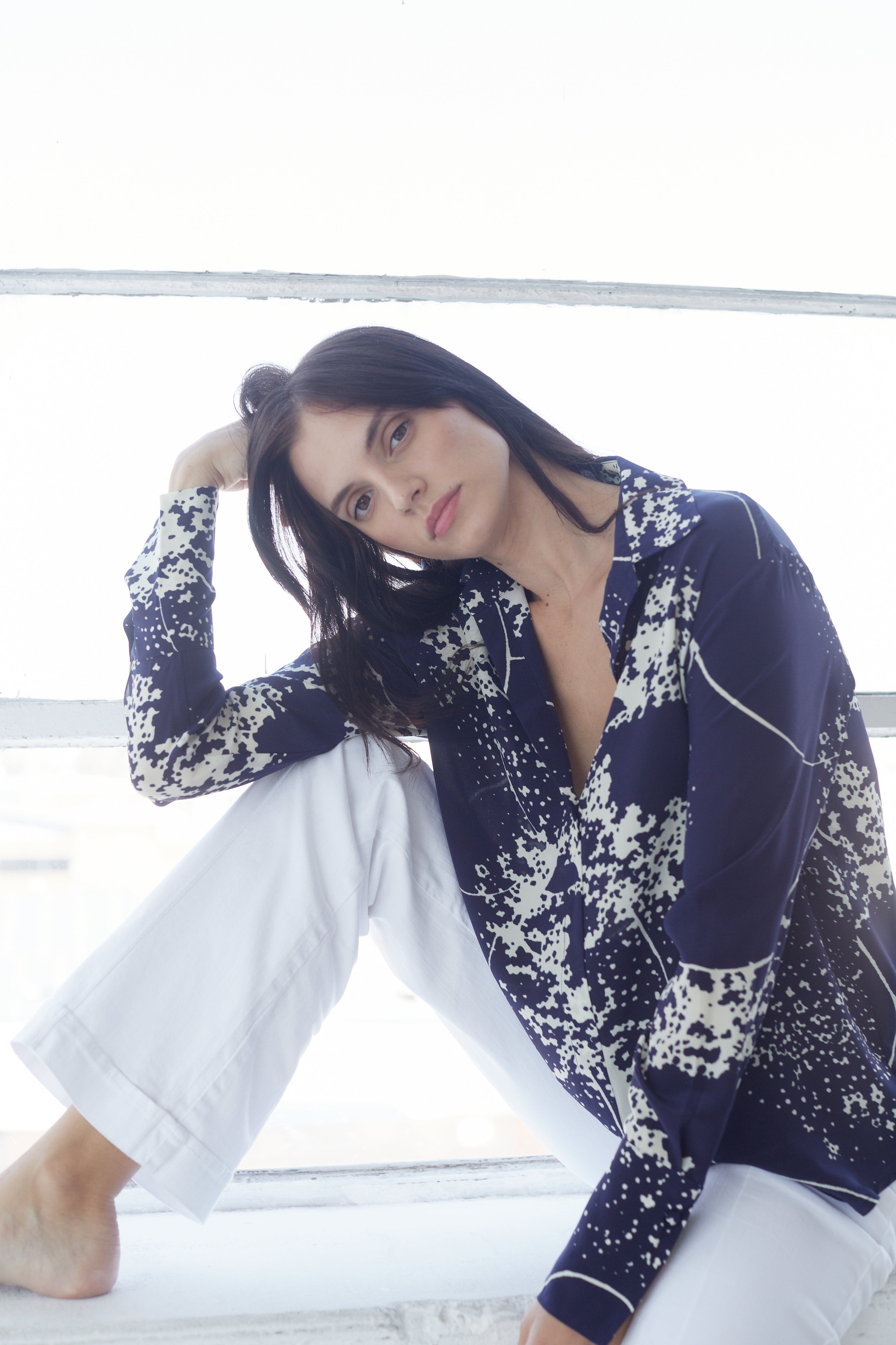 A woman sitting by a window wearing a Catherine Gee Daria French Cuff Silk Blouse - Cosmos Print features a dark blue background with white abstract patterns. She is also wearing white pants and is barefoot, with one leg bent and an arm resting on the knee.