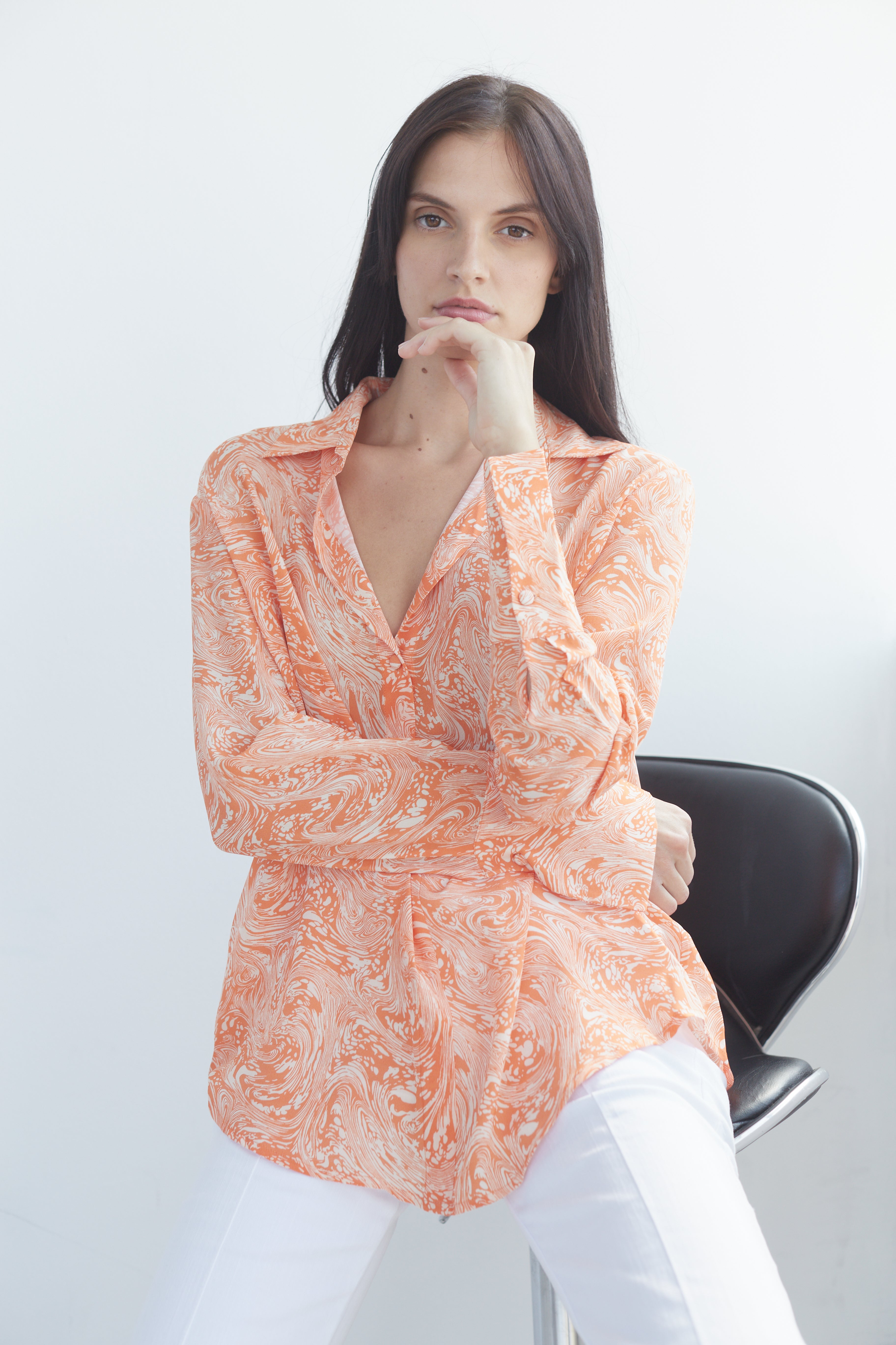A woman seated on a black stool, with arms crossed and one hand resting near the face wearing a Catherine Gee Daria French Cuff Silk Sorbet Swirl blouse that features a vibrant orange and white swirl pattern, long sleeves with French cuffs, and a relaxed fit. The blouse's elegant design and intricate pattern make it a standout piece, highlighting both style and sophistication.