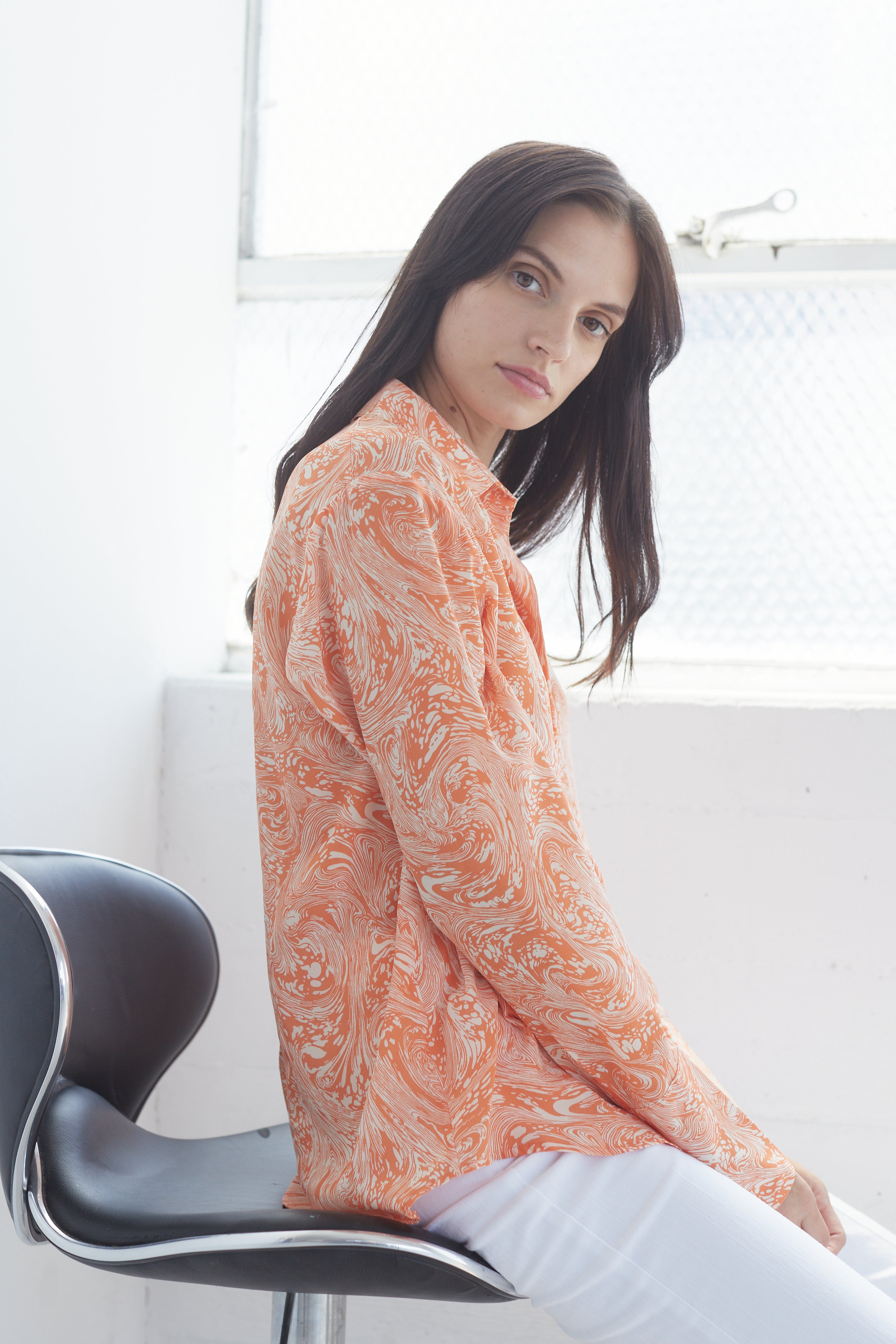 Women seated on a modern black chair, wearing a Catherine Gee Daria French Cuff Silk Sorbet Swirl blouse features a vibrant orange and white swirl pattern, long sleeves, and a relaxed fit paired with white pants. The setting appears to be a bright room with a large window in the background, allowing natural light to illuminate the scene. The blouse's intricate design and luxurious silk fabric make it a standout piece, highlighting its elegance and style.