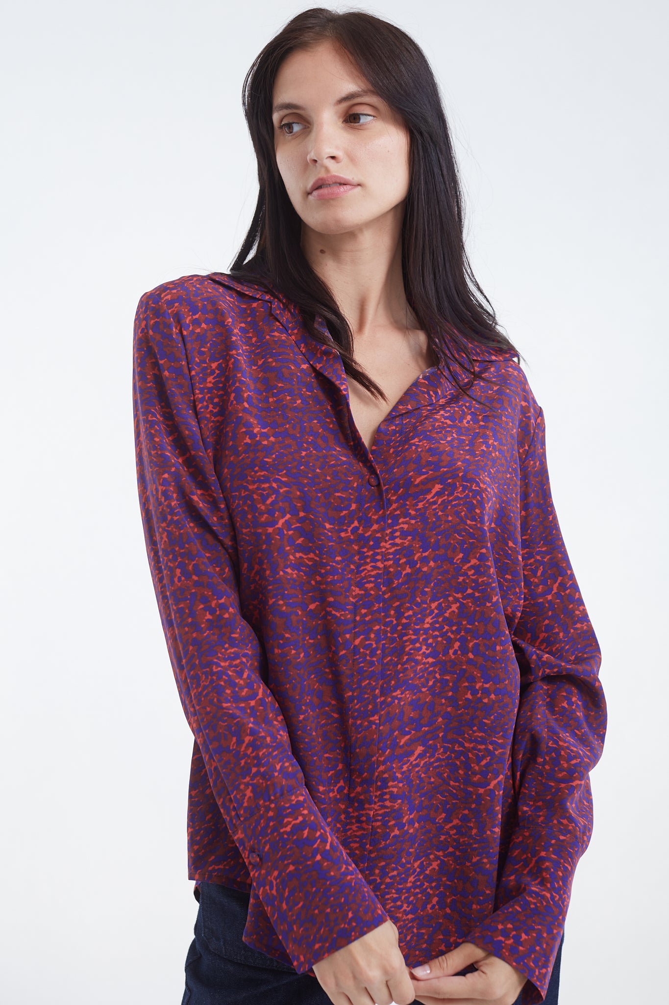 A woman standing with hands clasped in front, on a plain white background, wearing a Catherine Gee Daria French Cuff Silk-Milan Print blouse features a vibrant pattern with a mix of red and purple colors. It has long sleeves with French cuffs and a V-neckline.