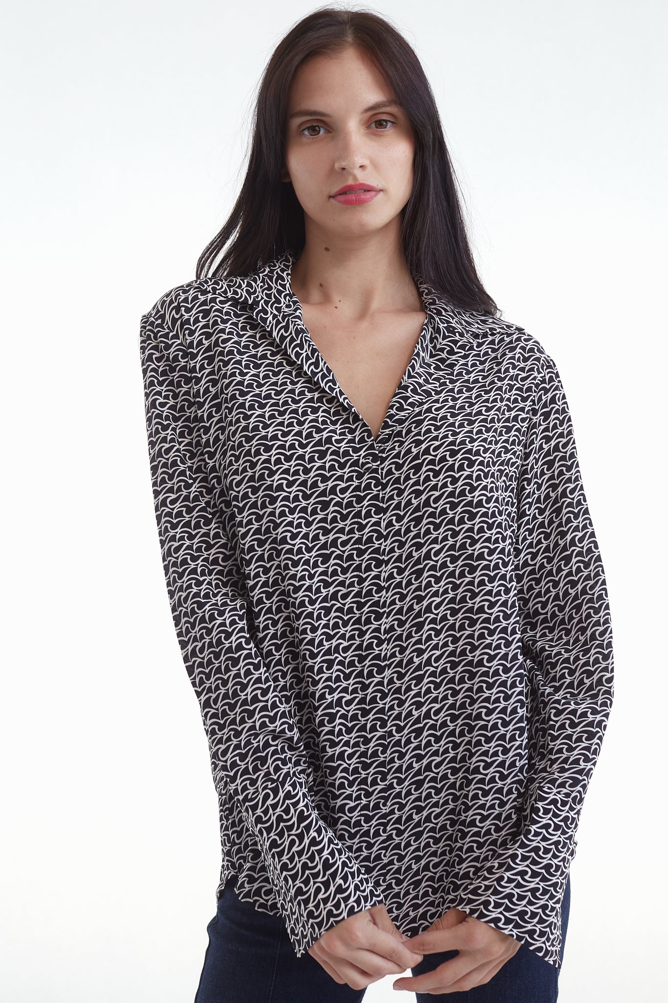 A woman wearing a Catherine Gee Daria French Cuff Silk-Florence Print blouse features a black and white pattern with a V-neck collar and long sleeves. She is standing with hands clasped in front on a plain white background.