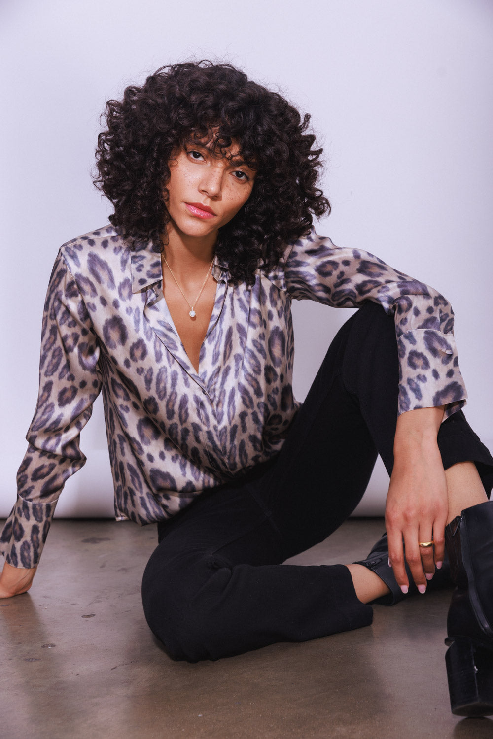 A woman sitting on the floor against a plain background wearing a Catherine Gee Daria French Cuff Silk New Leopard Blouse features a leopard print design, a silky texture, and a loose fit, with French cuffs on the sleeves paired with black pants and black boots. It highlights the stylish and elegant design of the blouse, making it interesting and relevant for fashion enthusiasts.