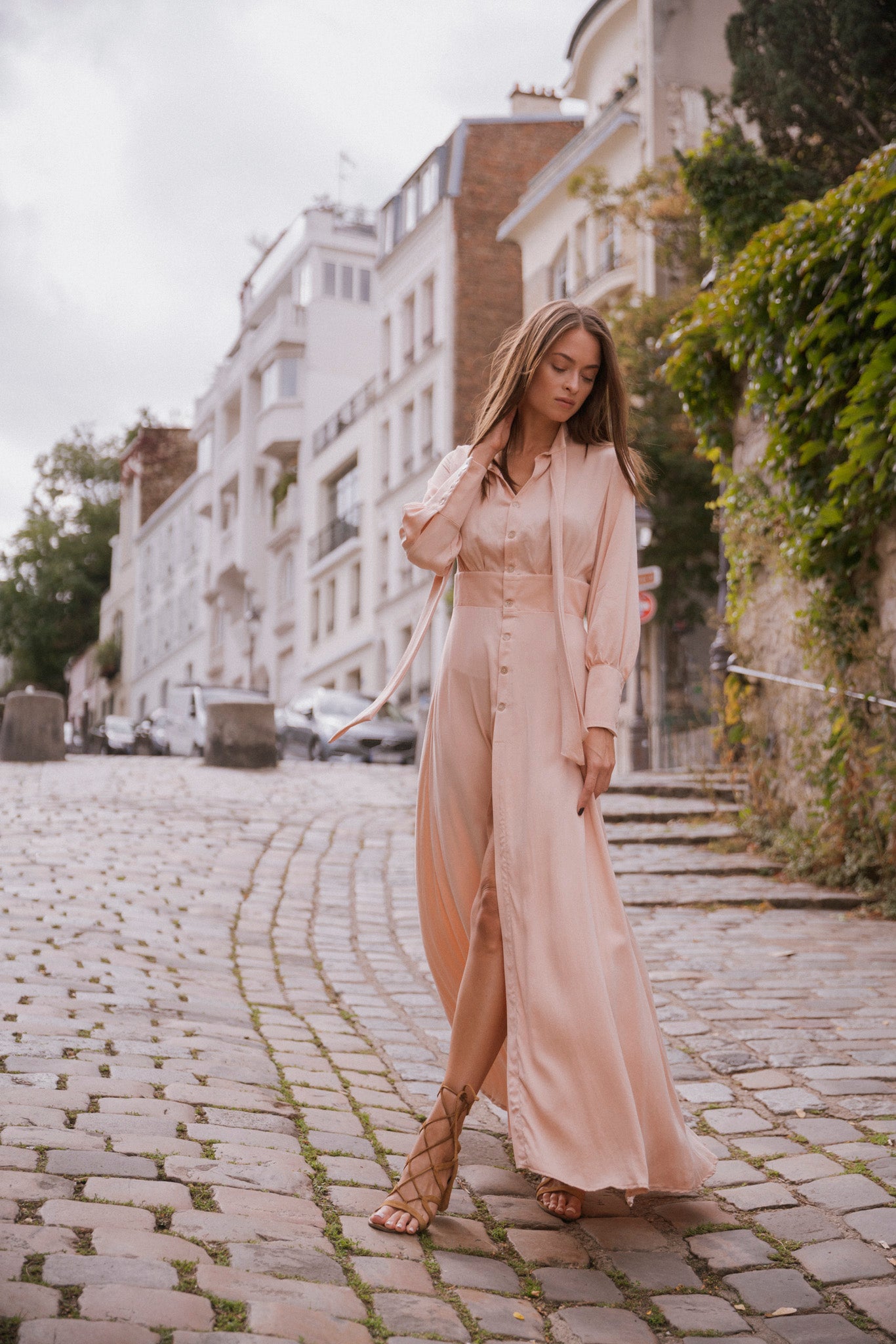 A woman wearing a Catherine Gee Electric Flou High-Slit Long Dress. This Nude color, long-sleeved dress features a high slit that reveals the legs, enhancing its elegant and flowing design paired with strappy sandals, creating a chic and sophisticated look. The backdrop includes a charming, slightly inclined cobblestone street with parked cars and greenery attached crawling to the buildings, adding a picturesque touch to the scene.