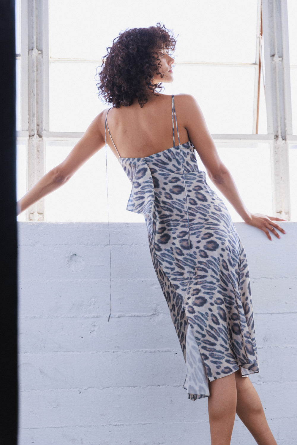 A woman standing against a white wall with large windows wearing the Catherine Gee Emma New Leopard Slip Dress allowing natural light to enhance the dress's elegant design. This dress features a chic leopard print pattern in shades of grey, black, and white. It has thin spaghetti straps and a fitted silhouette that flares slightly at the hem, creating a sophisticated and stylish look that highlights the dress's luxurious fabric and trendy pattern.