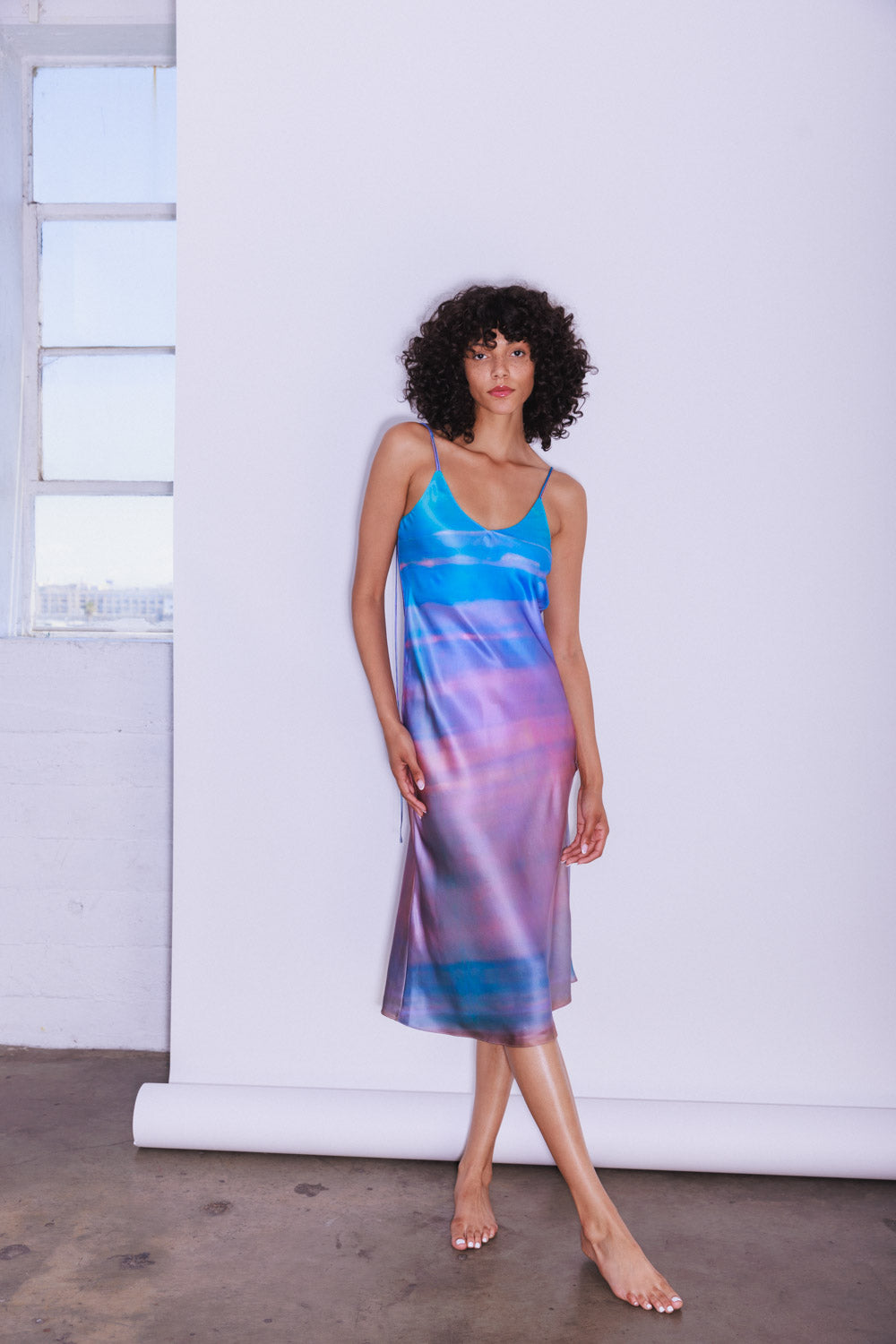 A woman wearing the Catherine Gee Emma Peruvian Quartz Slip dress features a vibrant, multicolored pattern with shades of blue, purple, and pink, resembling a watercolor effect. It has thin spaghetti straps and a V-neckline, falling to a midi length. The minimalist studio setting, with a white backdrop and a window allowing natural light, highlights the dress's luxurious fabric and striking design.