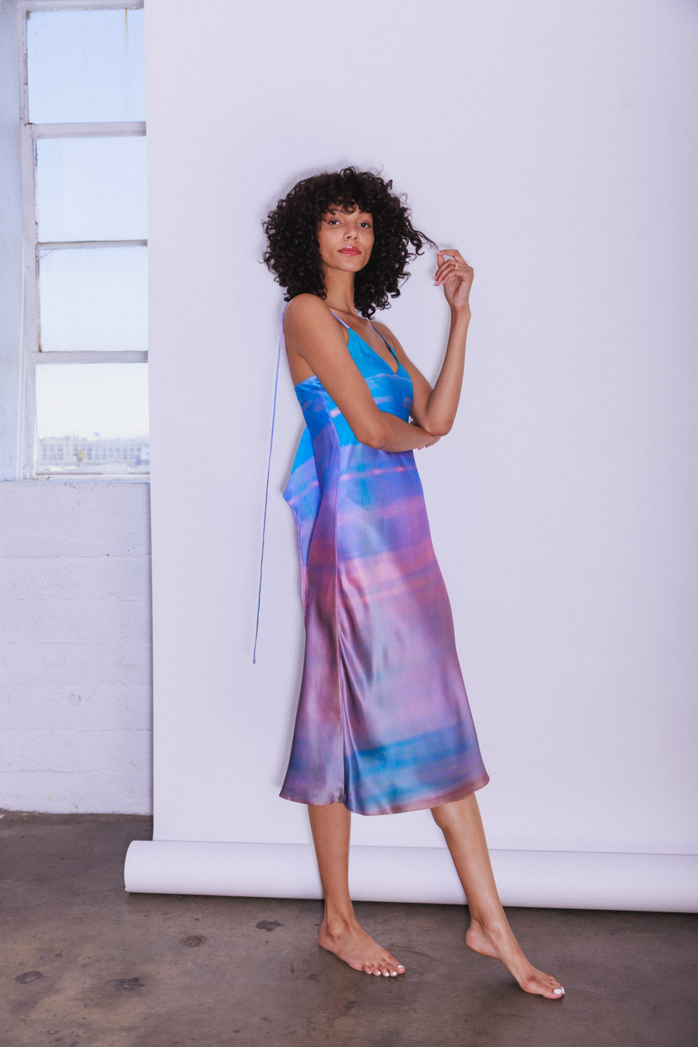 A woman wearing a Catherine Gee Emma Peruvian Quartz Slip dress stands barefoot on a concrete floor, with a white backdrop and window enhancing the dress's striking design. This elegant dress features a vibrant, multicolored pattern in shades of blue, purple, and pink, reminiscent of Peruvian quartz. It has thin spaghetti straps and a flowing, midi-length silhouette that drapes gracefully. The dress is made of silky charmeuse fabric, giving it a luxurious and fluid appearance.