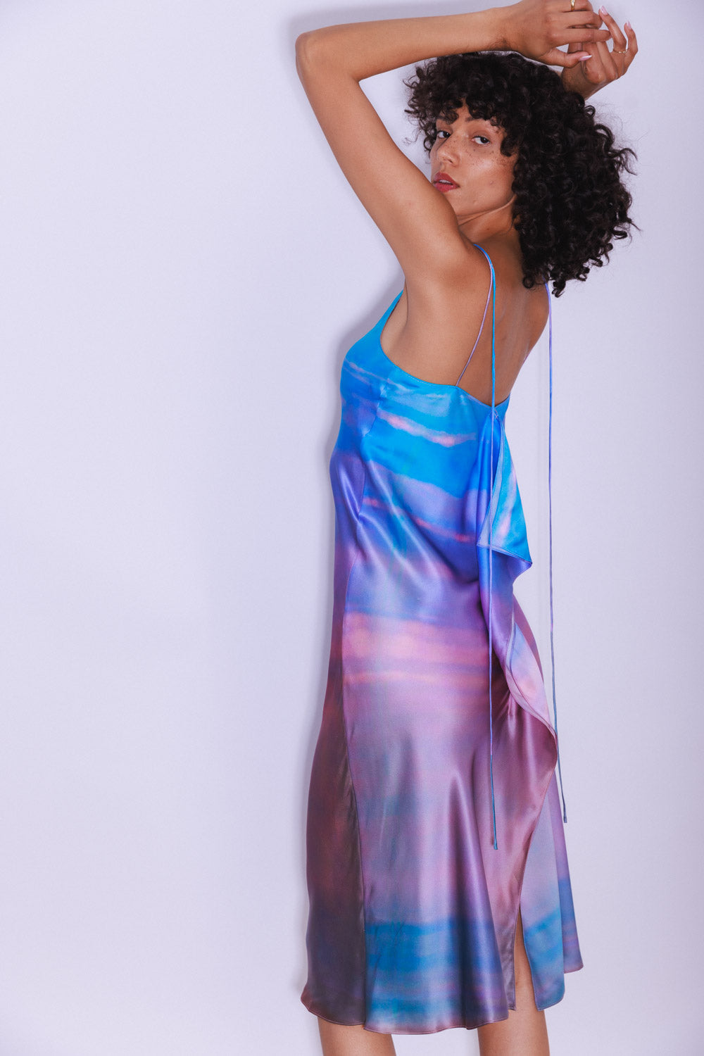 A woman posing with her arms raised wearing the Catherine Gee Emma Peruvian Quartz Slip dress features a vibrant, multicolored design with shades of blue, purple, and pink, resembling a watercolor effect. It has thin spaghetti straps and a loose, flowing fit that drapes elegantly. The back of the dress has a cascading ruffle detail, adding a touch of sophistication and highlighting the dress's fluidity and style.